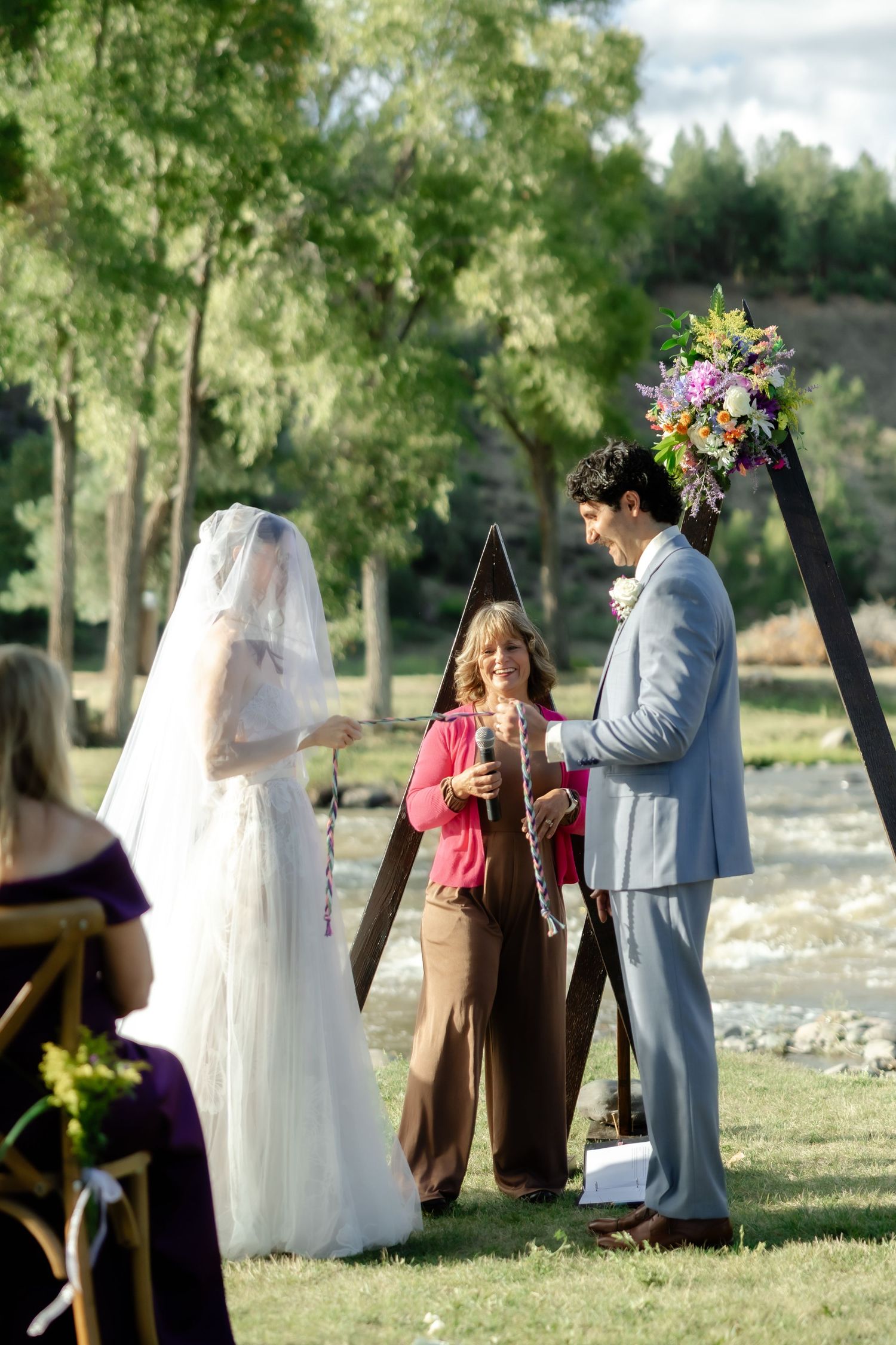 Bride and groom exchange vows outdoors with officiant at a wooden arch decorated with colorful flowers.