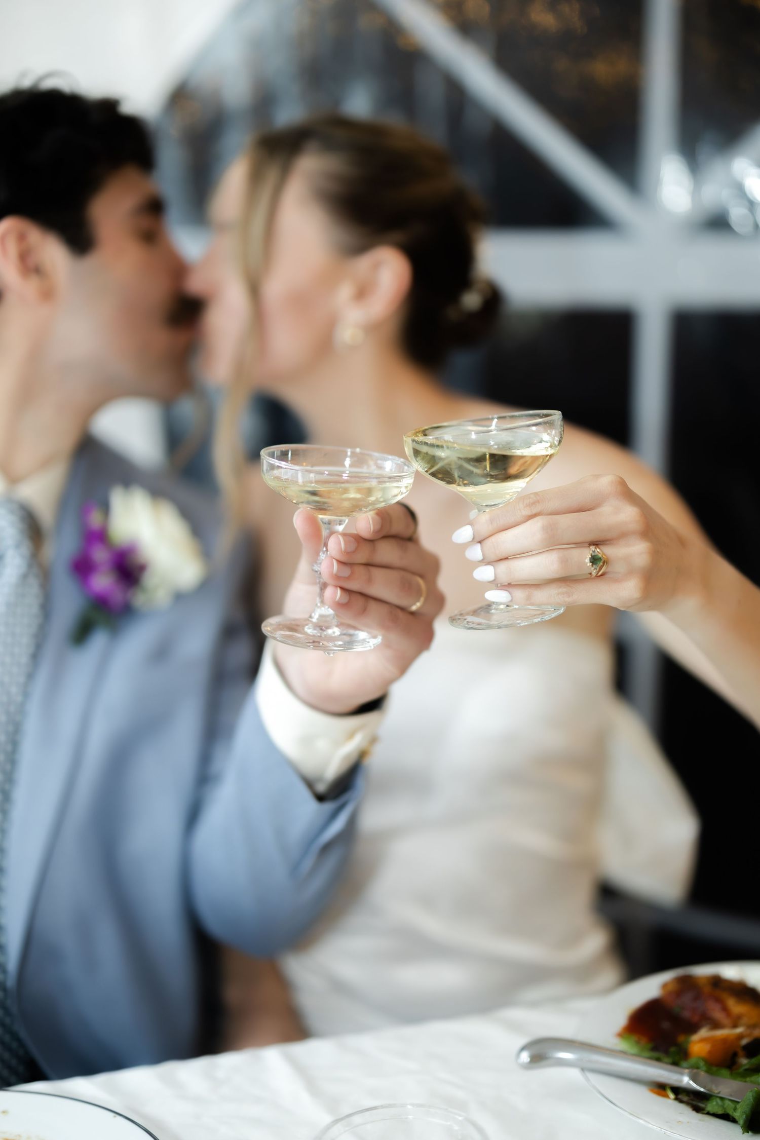 Newlywed couple kissing while clinking champagne glasses at wedding reception table.