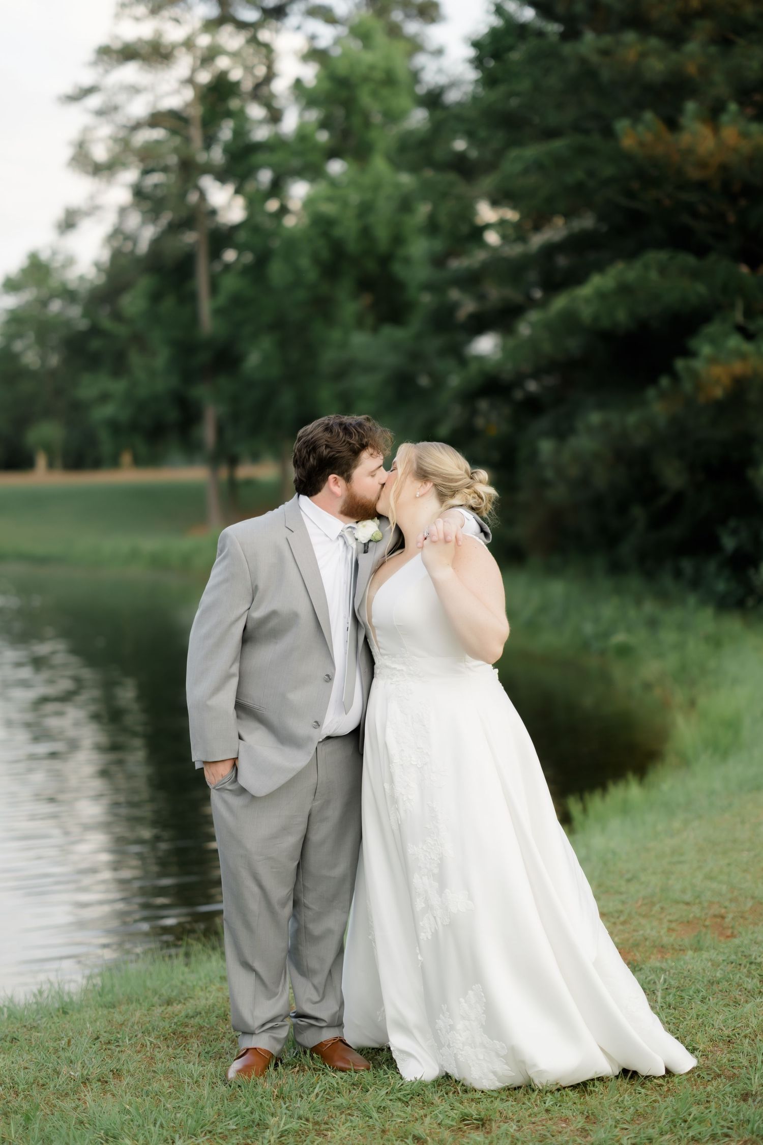 Bride and groom share a romantic kiss by a pond, dressed in a white gown and gray suit, surrounded by lush greenery.