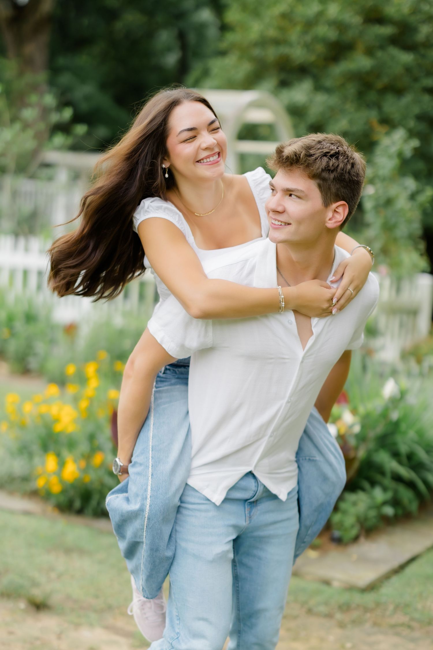 A young couple laughing outdoors, man giving woman a piggyback ride in a garden with colorful flowers.