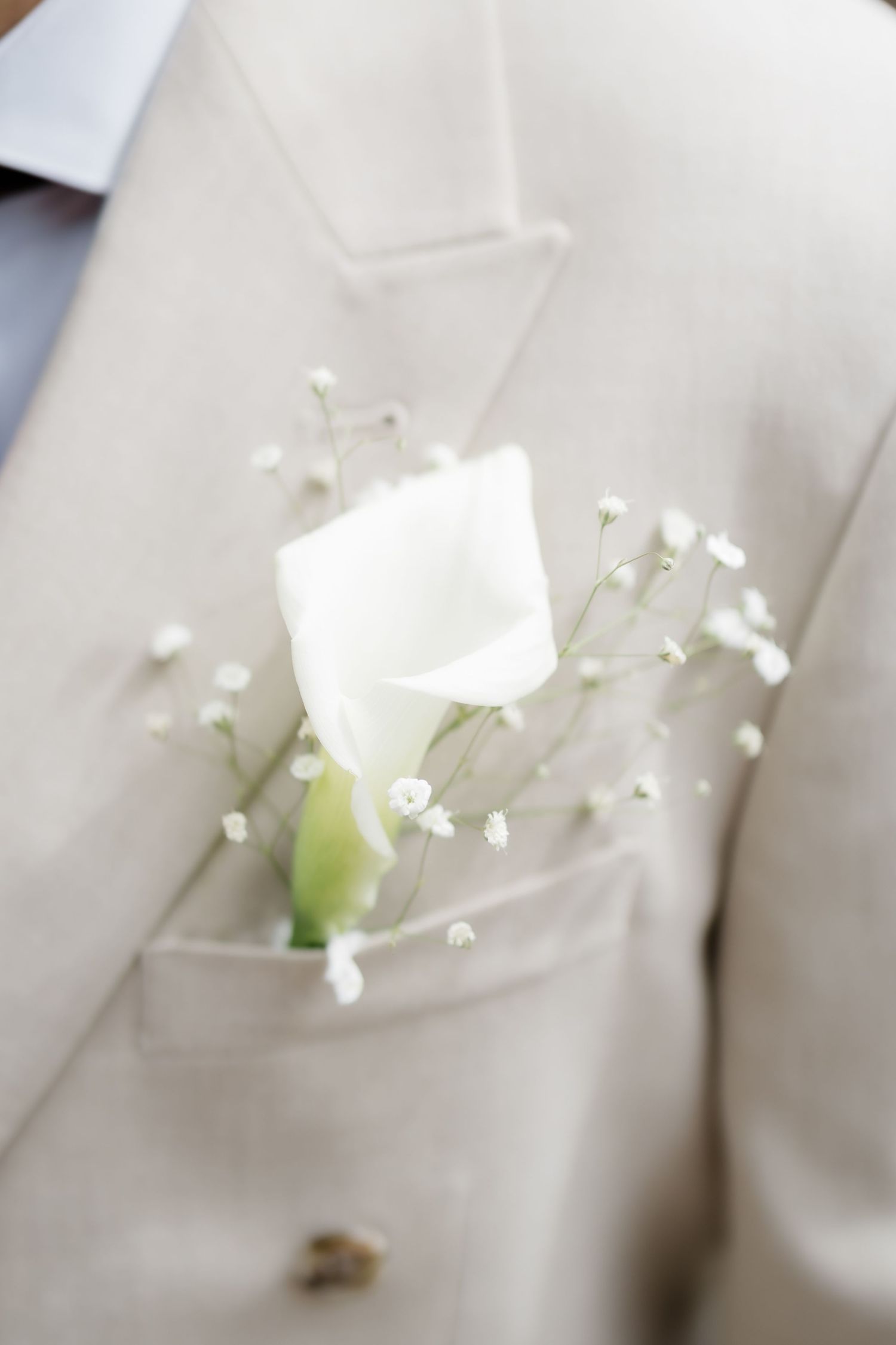 White calla lily and baby's breath boutonniere on a beige wedding suit lapel pocket.