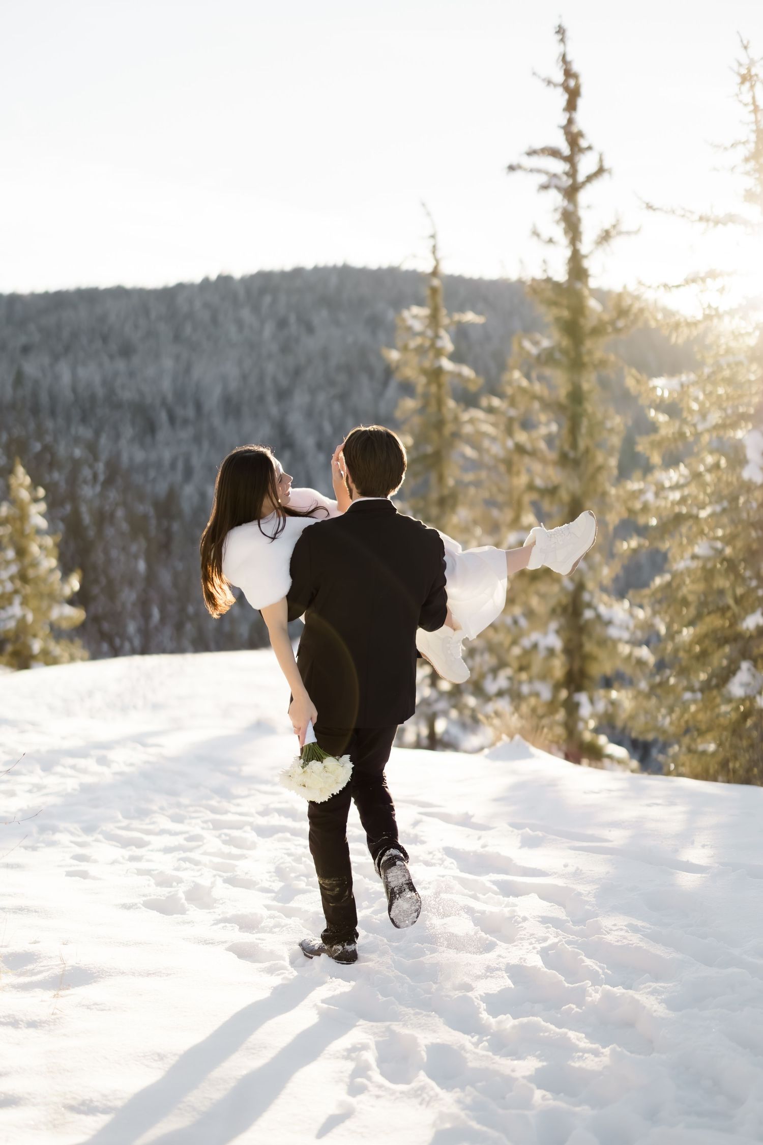 Groom carries bride in his arms while walking through snow with pine trees and mountains behind.