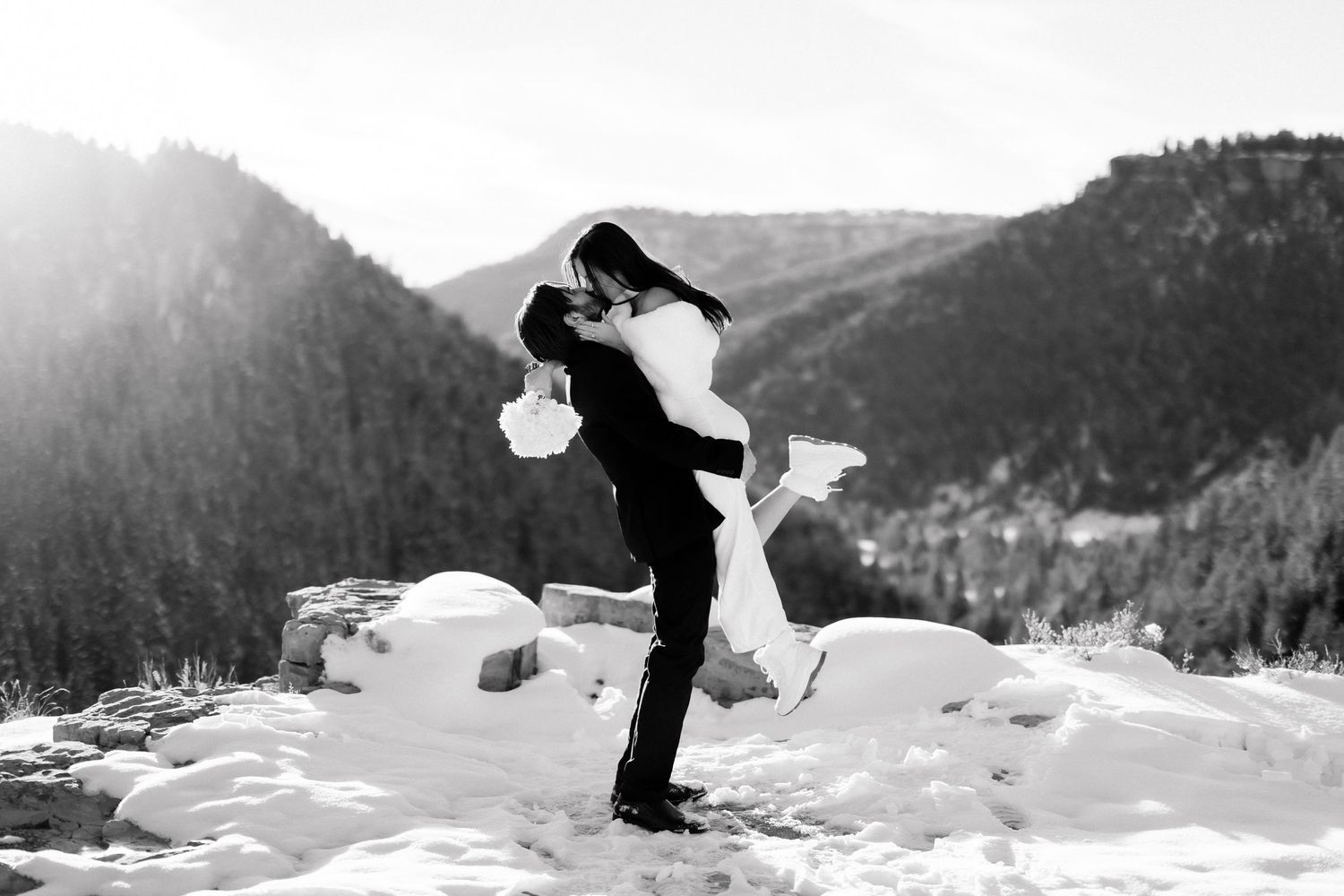 Black and white photo of groom lifting bride high in snowy mountain setting with bright sunlight.