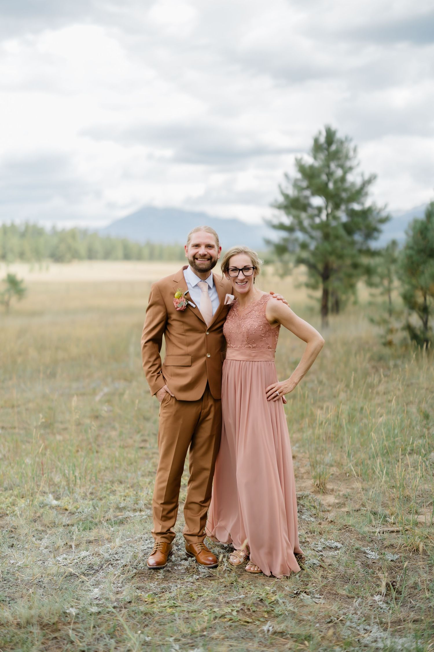 Couple posing in rustic mountain meadow, man in brown suit and woman in dusty rose gown, pine trees and mountains behind.