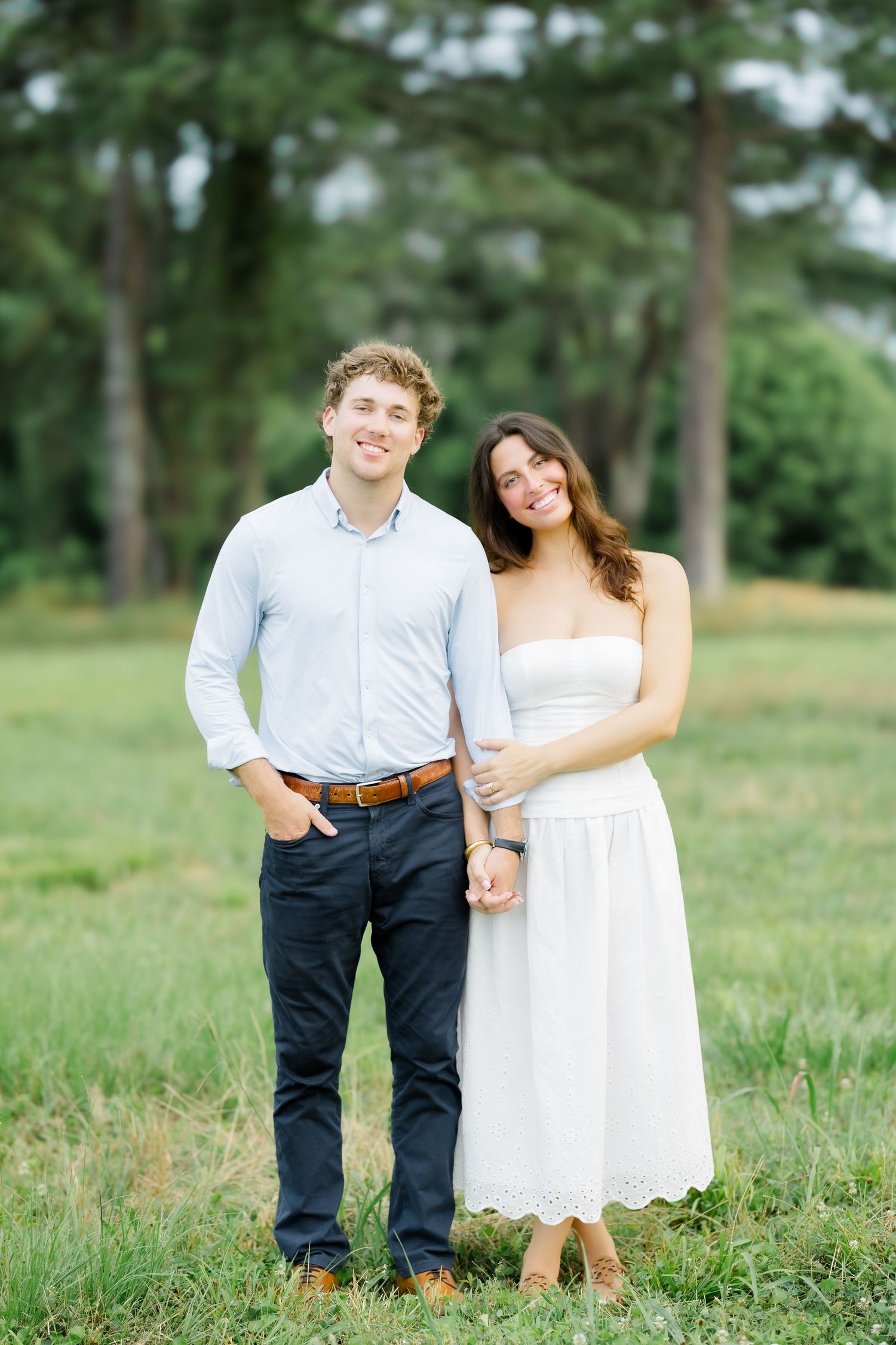A smiling couple poses in a green field, man in light blue shirt and dark jeans, woman in white strapless midi dress.