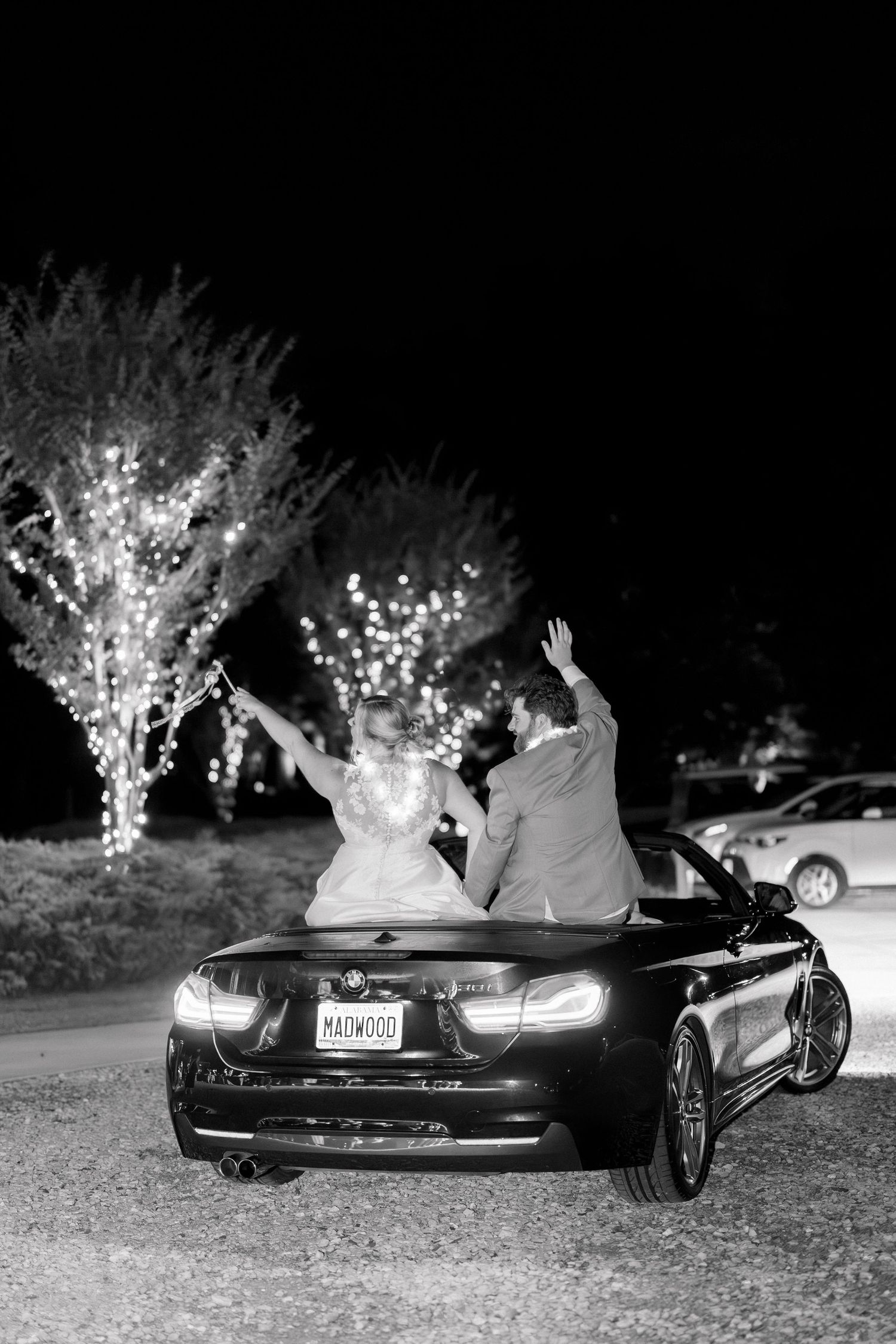 Bride and groom celebrate in a convertible BMW at night, arms raised, surrounded by twinkling lights.
