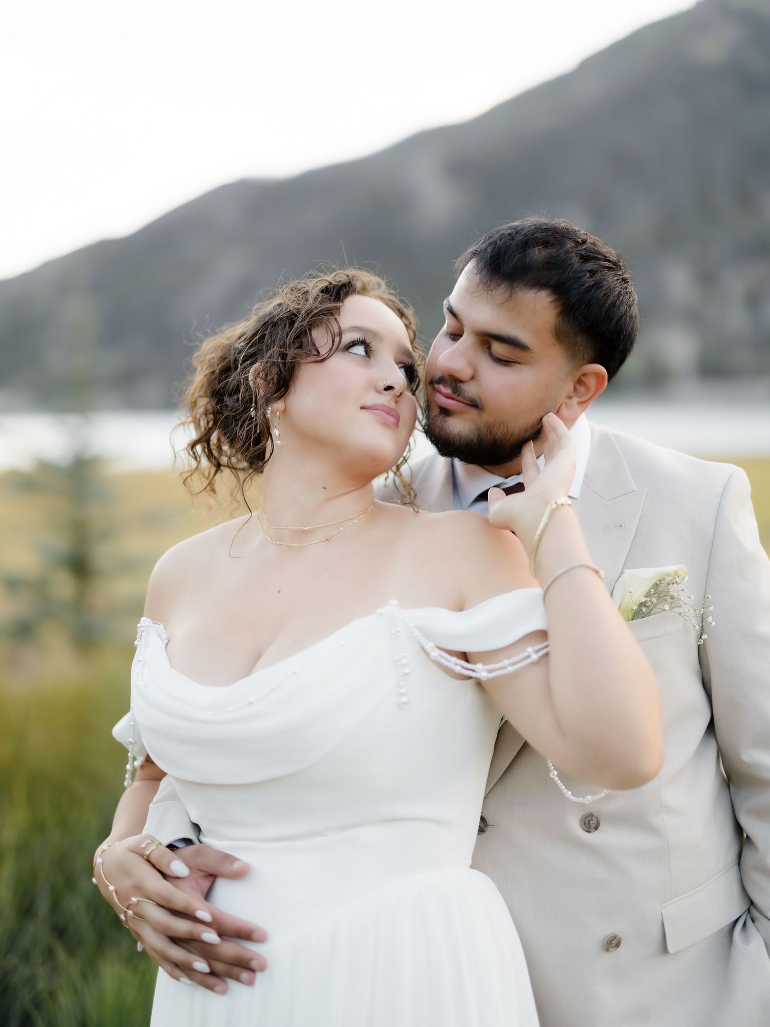 A bride in a white off-shoulder gown and groom in a beige suit share a romantic moment with mountains behind them.