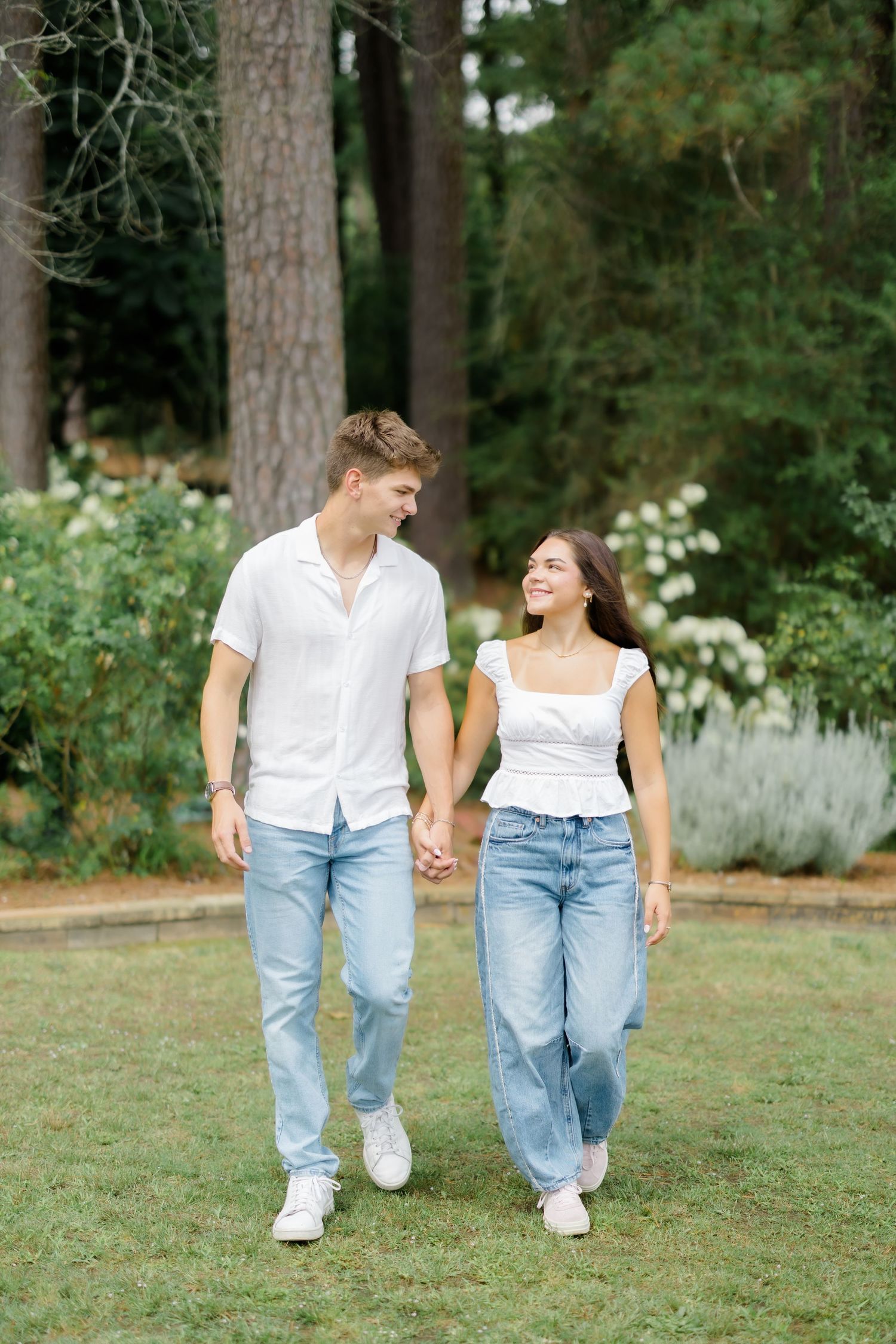 A happy young couple holding hands and walking together in a lush green garden wearing matching white tops and blue jeans.