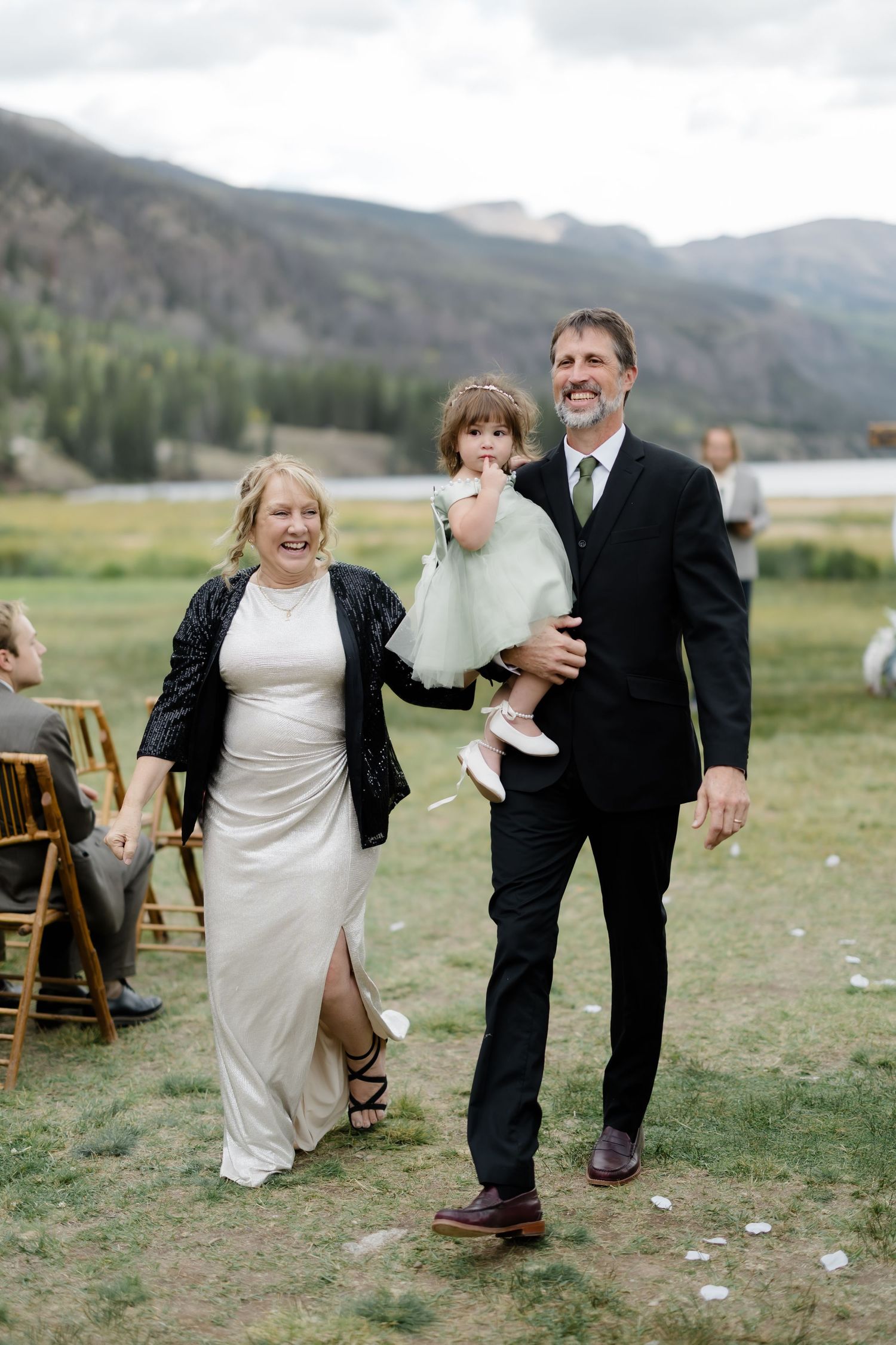 Smiling bride and groom walking down the aisle holding a young girl in a mint dress at an outdoor mountain wedding.
