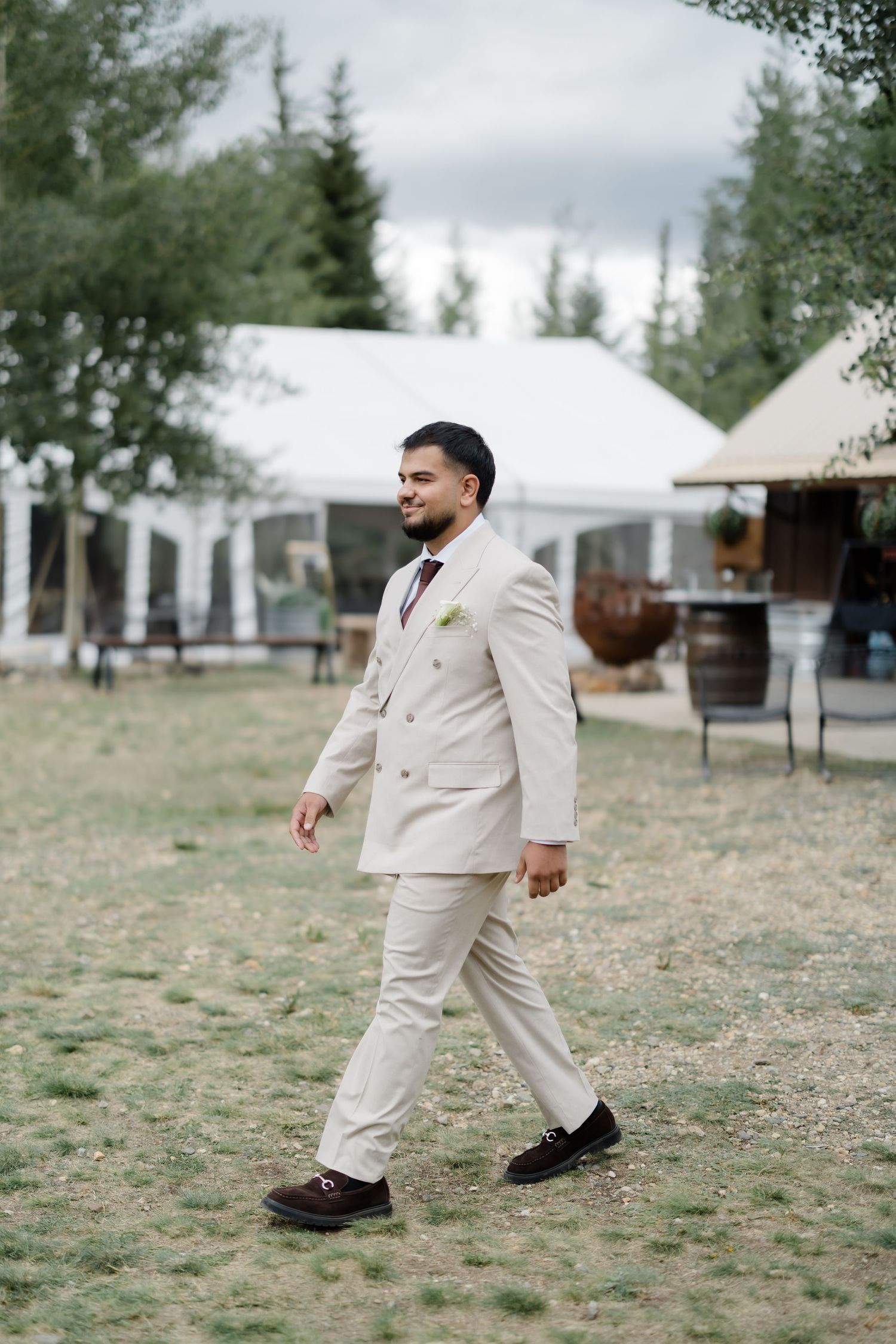 Groom in beige double-breasted suit walks outdoors at wedding venue with tents and trees in background.
