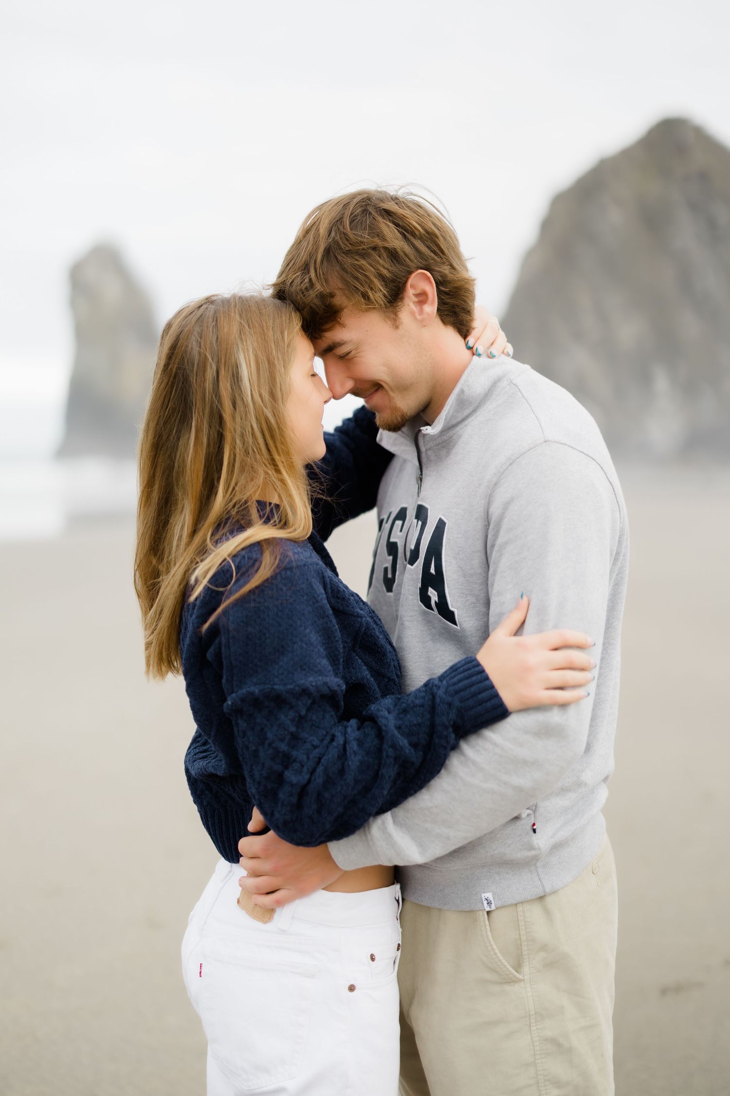 Couple embracing on a beach, touching foreheads romantically, with rocky sea stacks in the misty background.