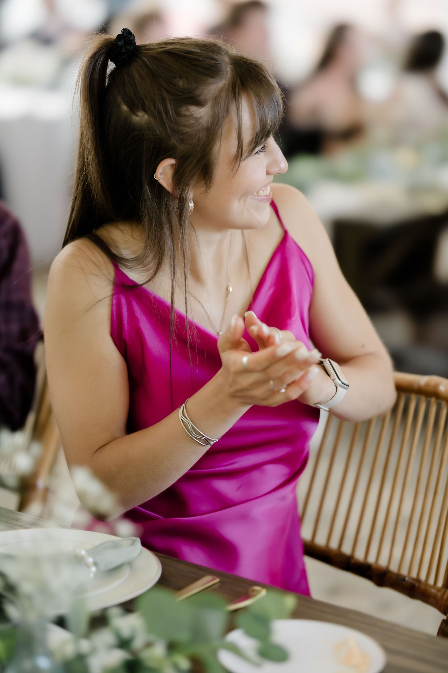 A smiling woman in a magenta satin slip dress claps her hands at an elegant wedding reception dinner.