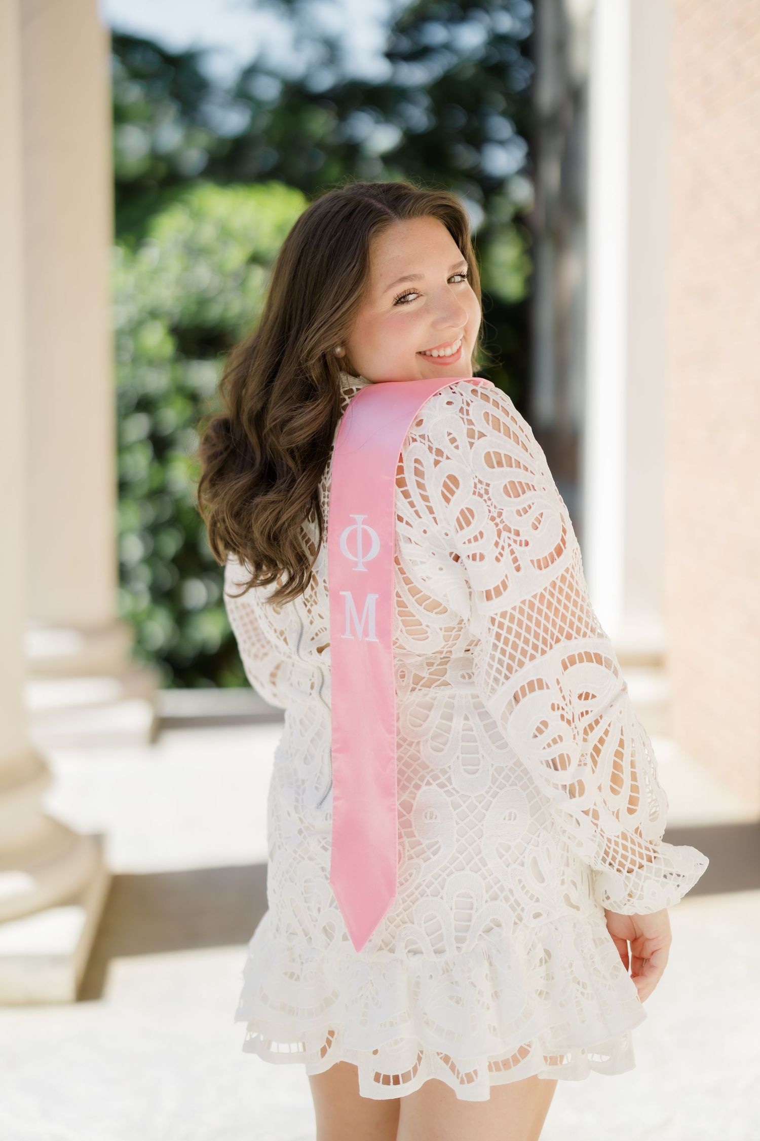 Young woman in white lace dress wearing pink Phi Mu sorority sash, smiling over shoulder outdoors.