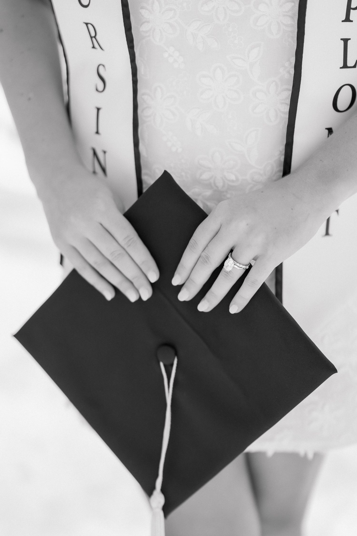Graduate holding a black mortarboard cap with tassel, wearing a white dress and sash, in a black and white photo.