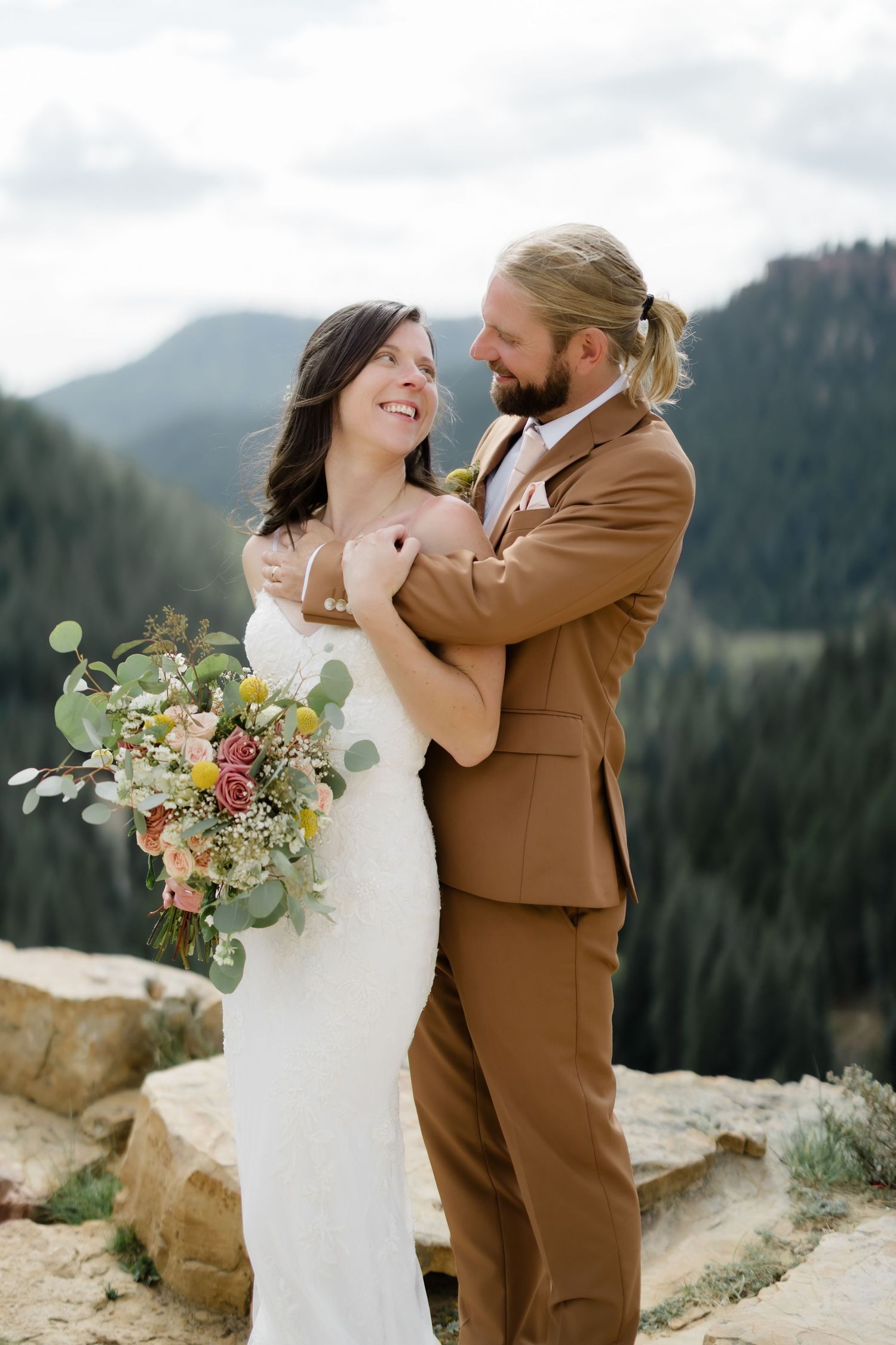 Happy bride in white lace gown and groom in tan suit embrace on mountain overlook with bouquet.