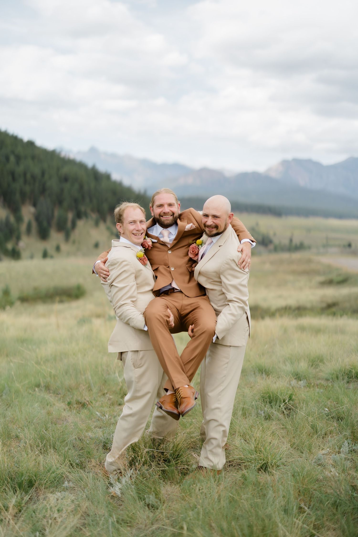 Two groomsmen in tan suits lift a smiling groom in a brown suit outdoors with mountain scenery behind them.