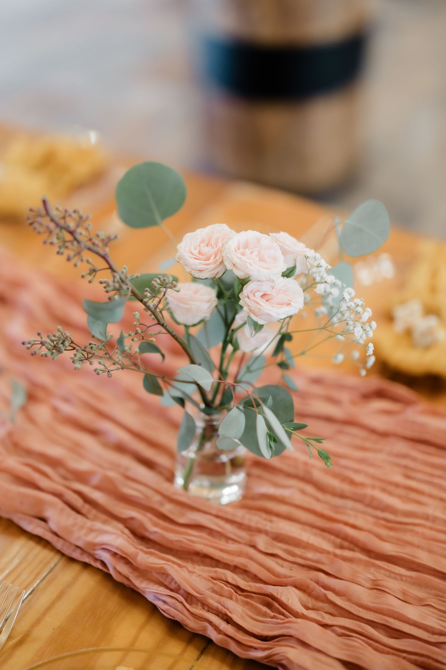 Small glass vase with blush pink roses and eucalyptus on a terracotta ruffled table runner atop a wooden table.