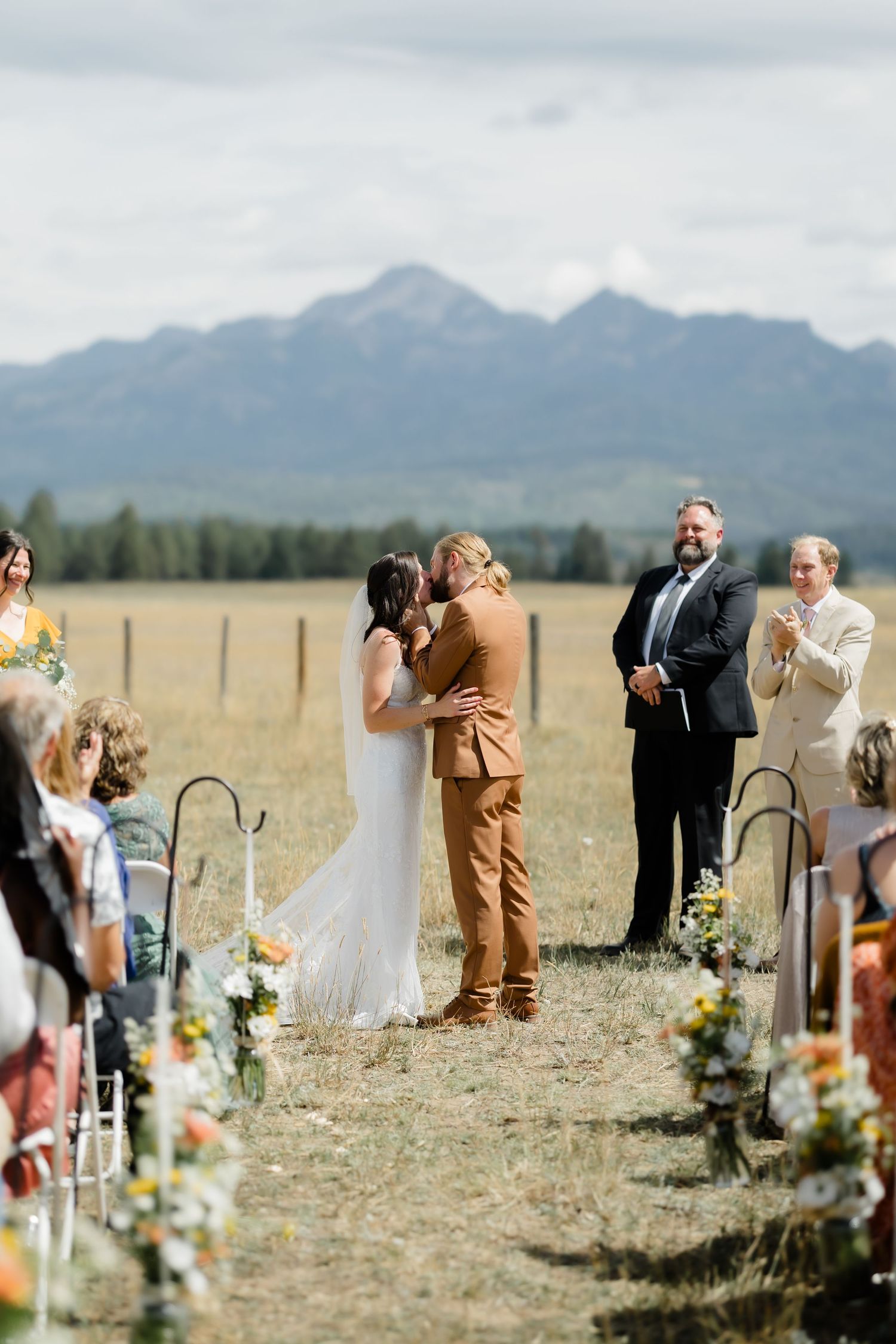 Bride and groom share first kiss at outdoor wedding ceremony with mountain backdrop, officiant and guests applauding nearby.
