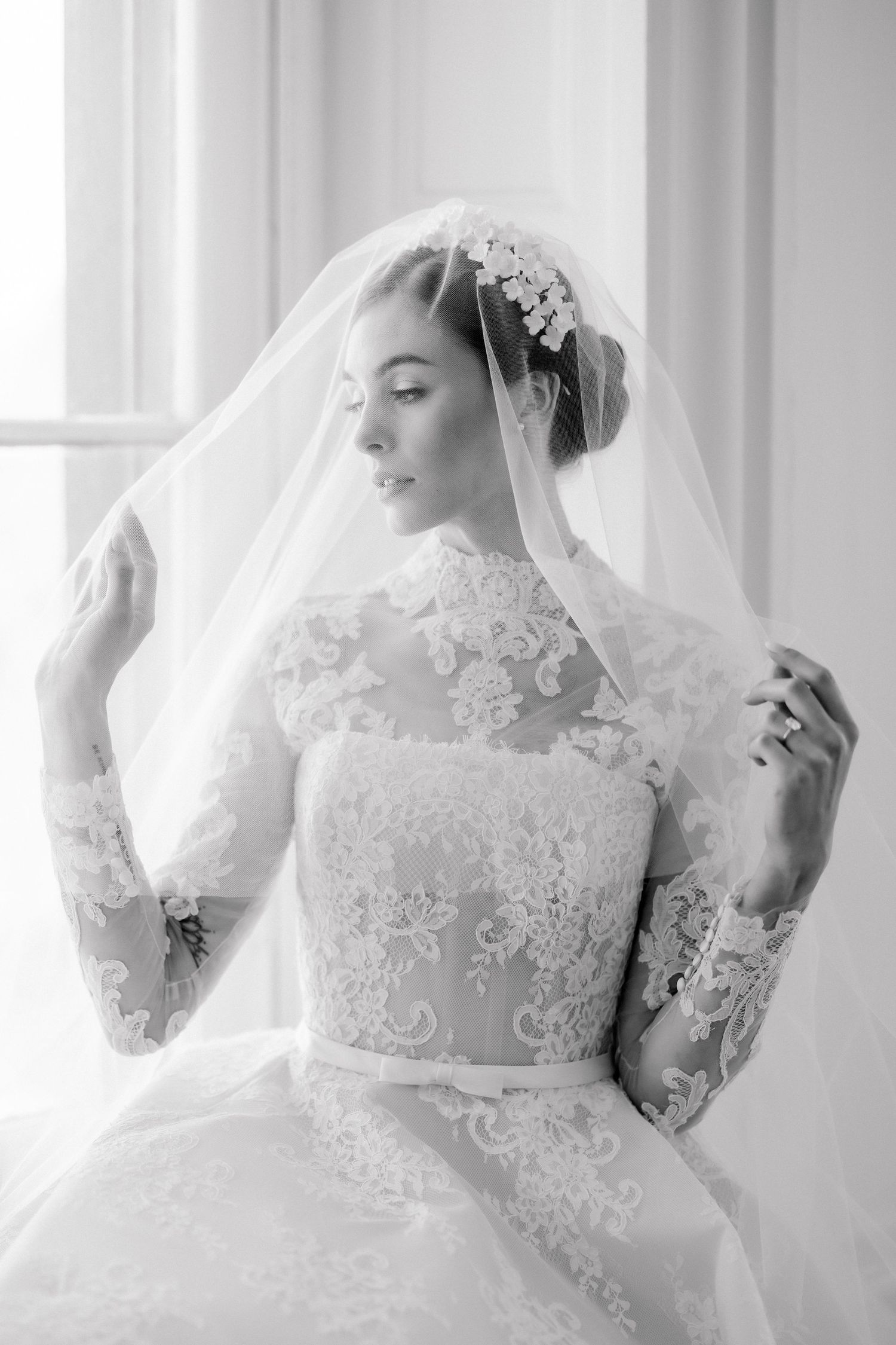 Black and white photo of a bride in lace wedding gown and veil, posing gracefully by a bright window.