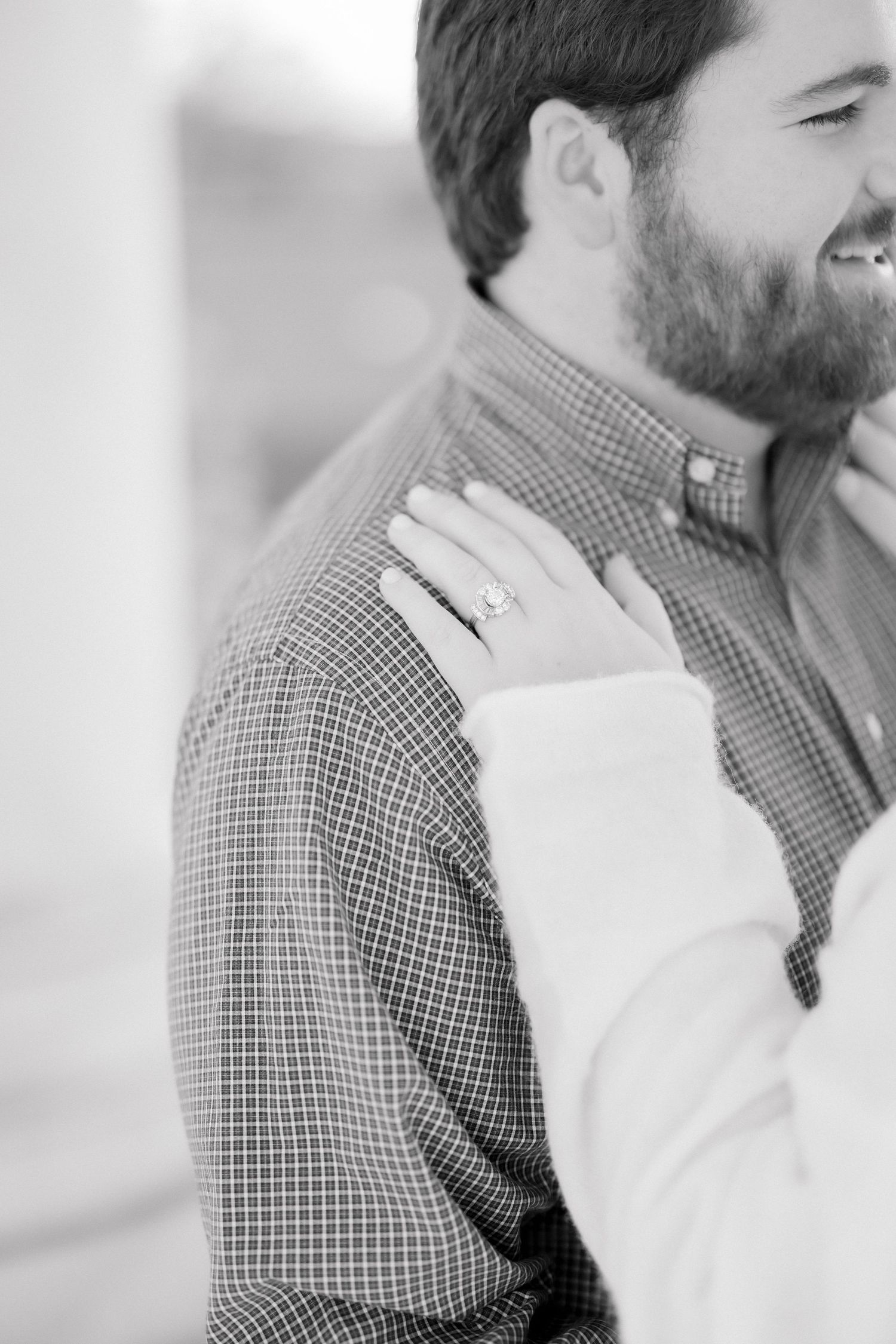 Close-up black and white photo of a woman's hand with engagement ring resting on man's shoulder.