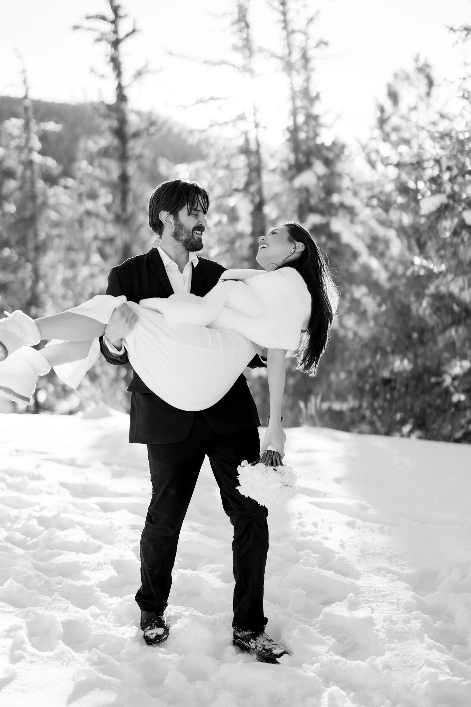Groom carries bride in snowy winter wonderland, black and white wedding photo with pine trees backdrop.