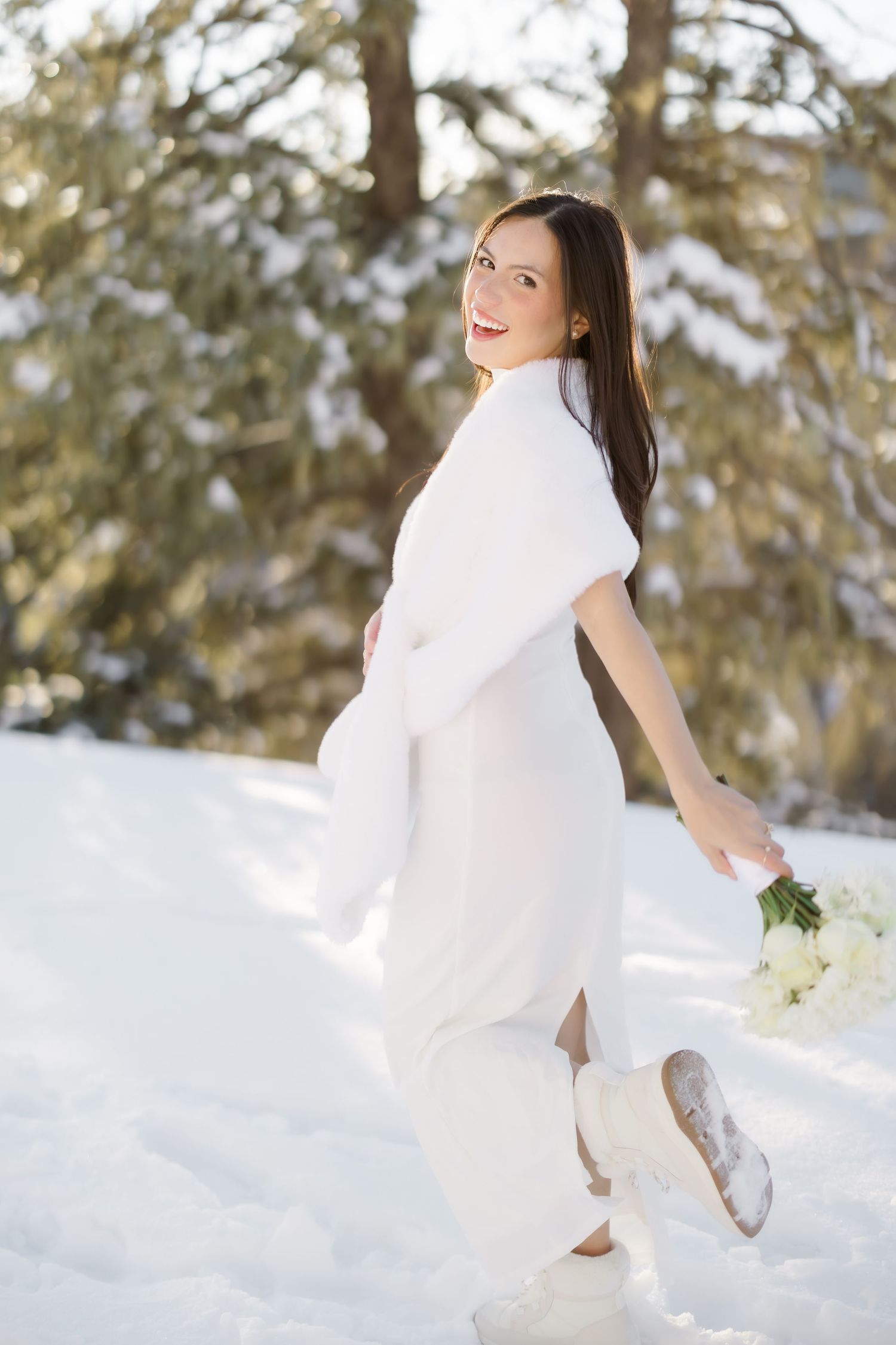 Smiling bride in white dress and fur wrap holds bouquet while walking through snowy winter landscape.