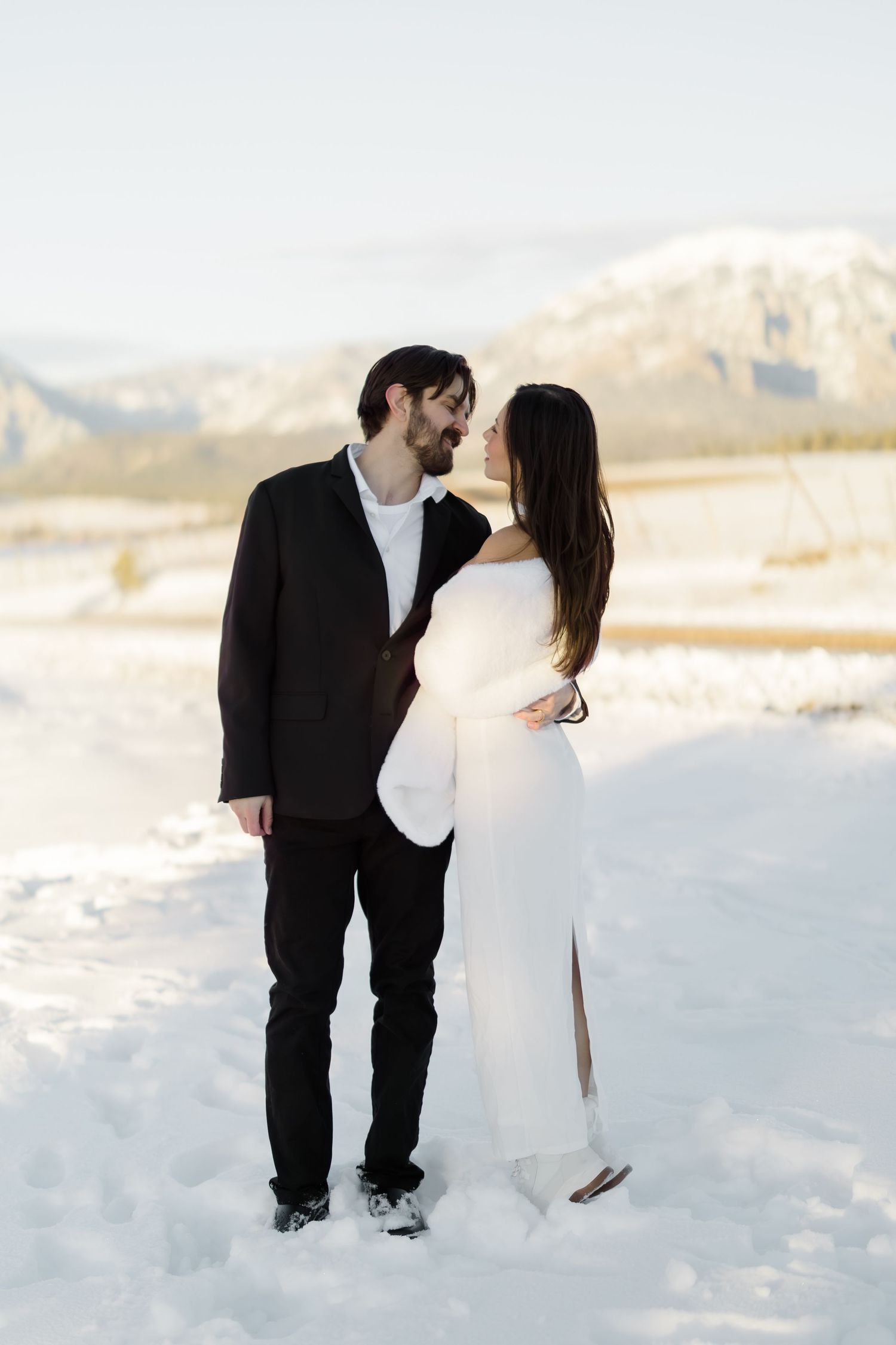 Bride and groom share a romantic moment standing close together in a snowy mountain setting.