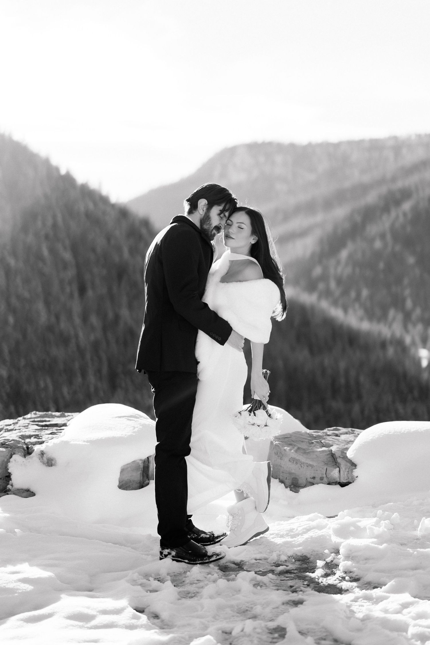 Black and white photo of bride and groom embracing on snowy mountain rocks with forested hills behind.