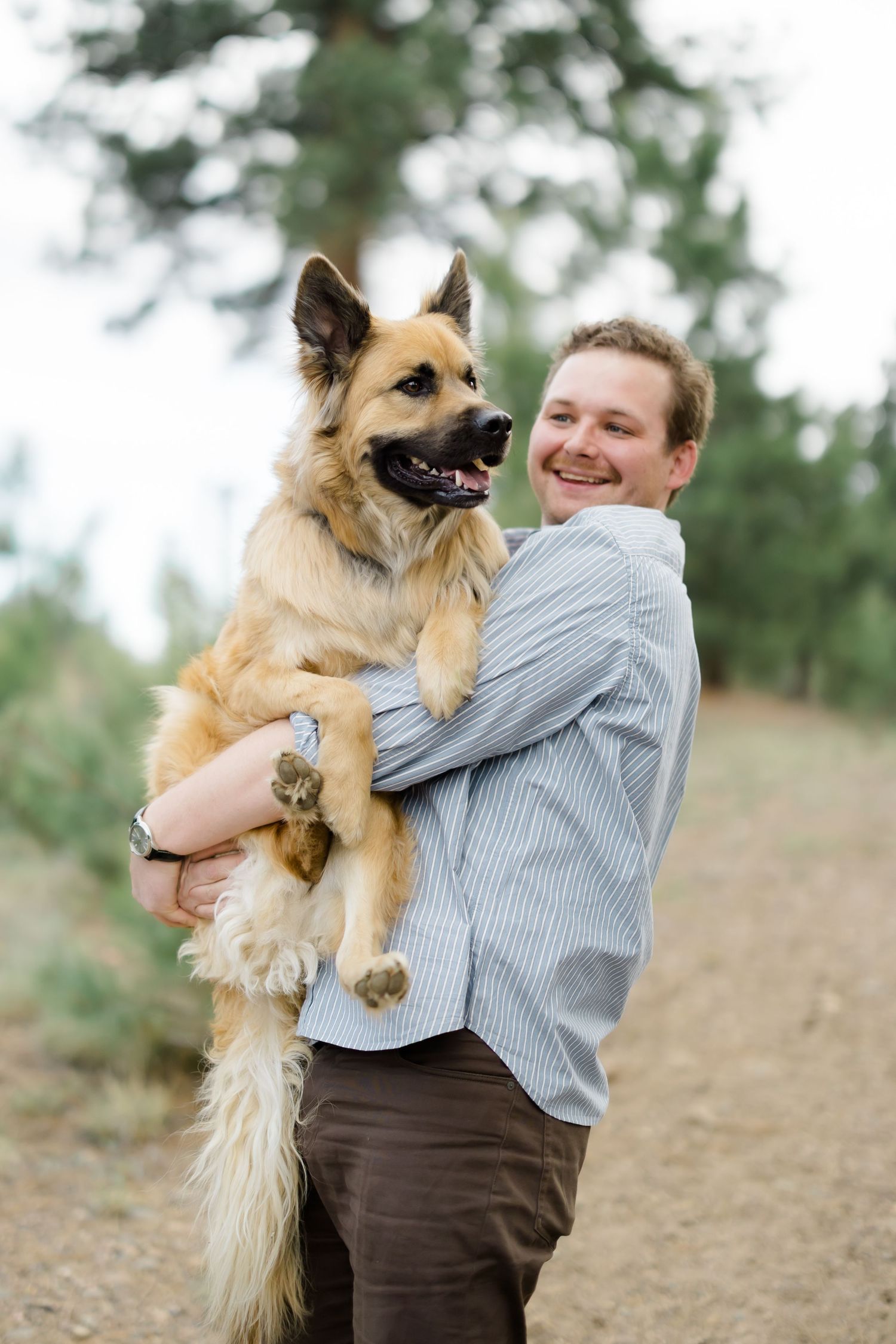 Smiling man holding a large German Shepherd dog on a forest trail outdoors.