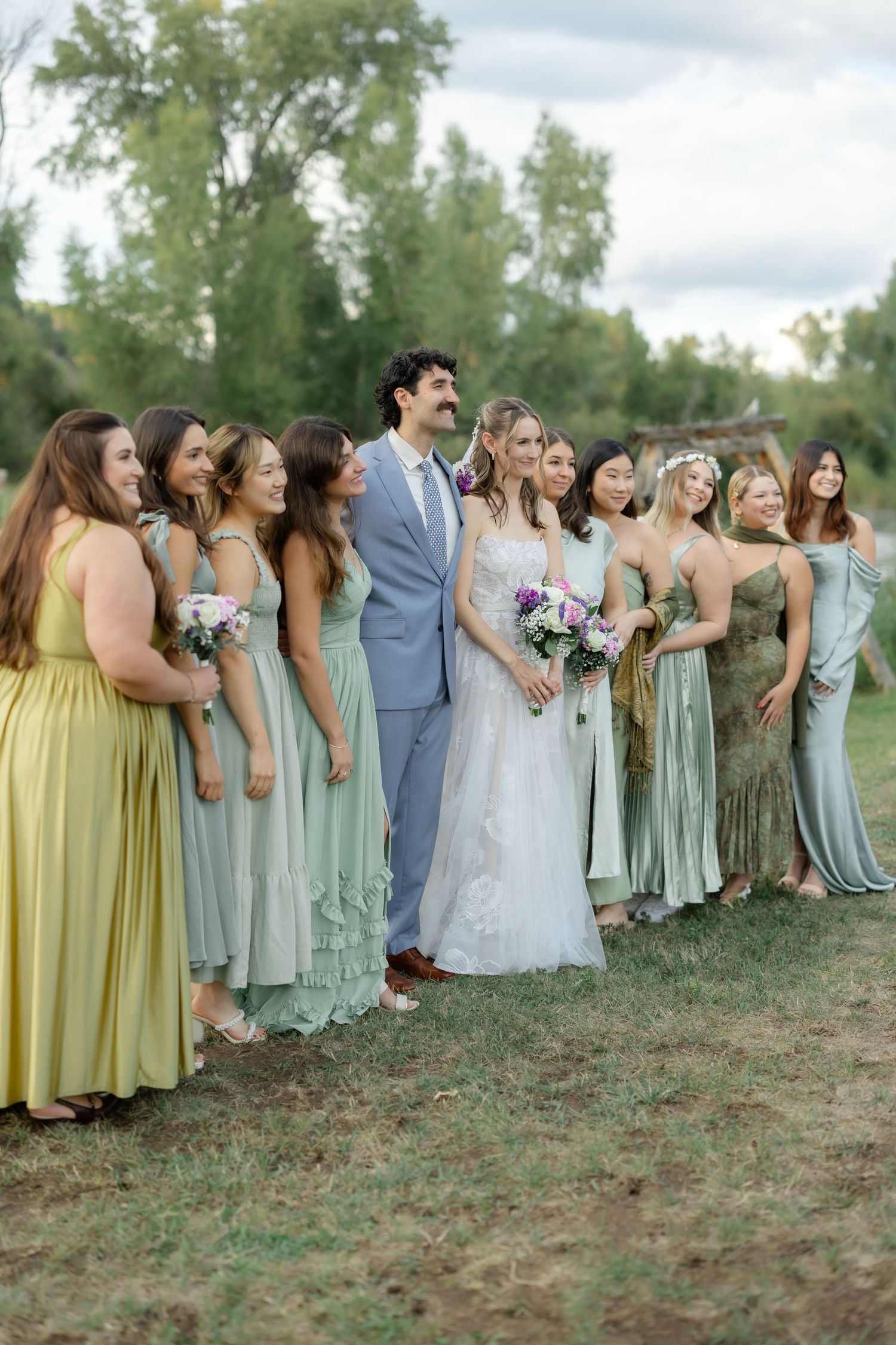 Bride and groom pose with bridesmaids in sage green and yellow gowns at an outdoor wedding ceremony.