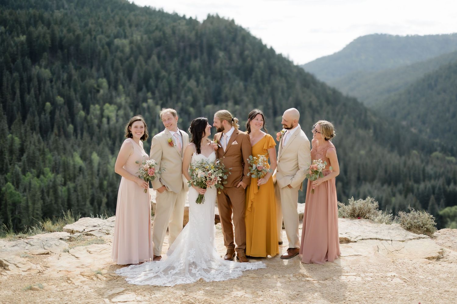 Wedding party poses on mountain overlook with pine forest backdrop, bride in white gown, bridesmaids in blush and mauve dresses.