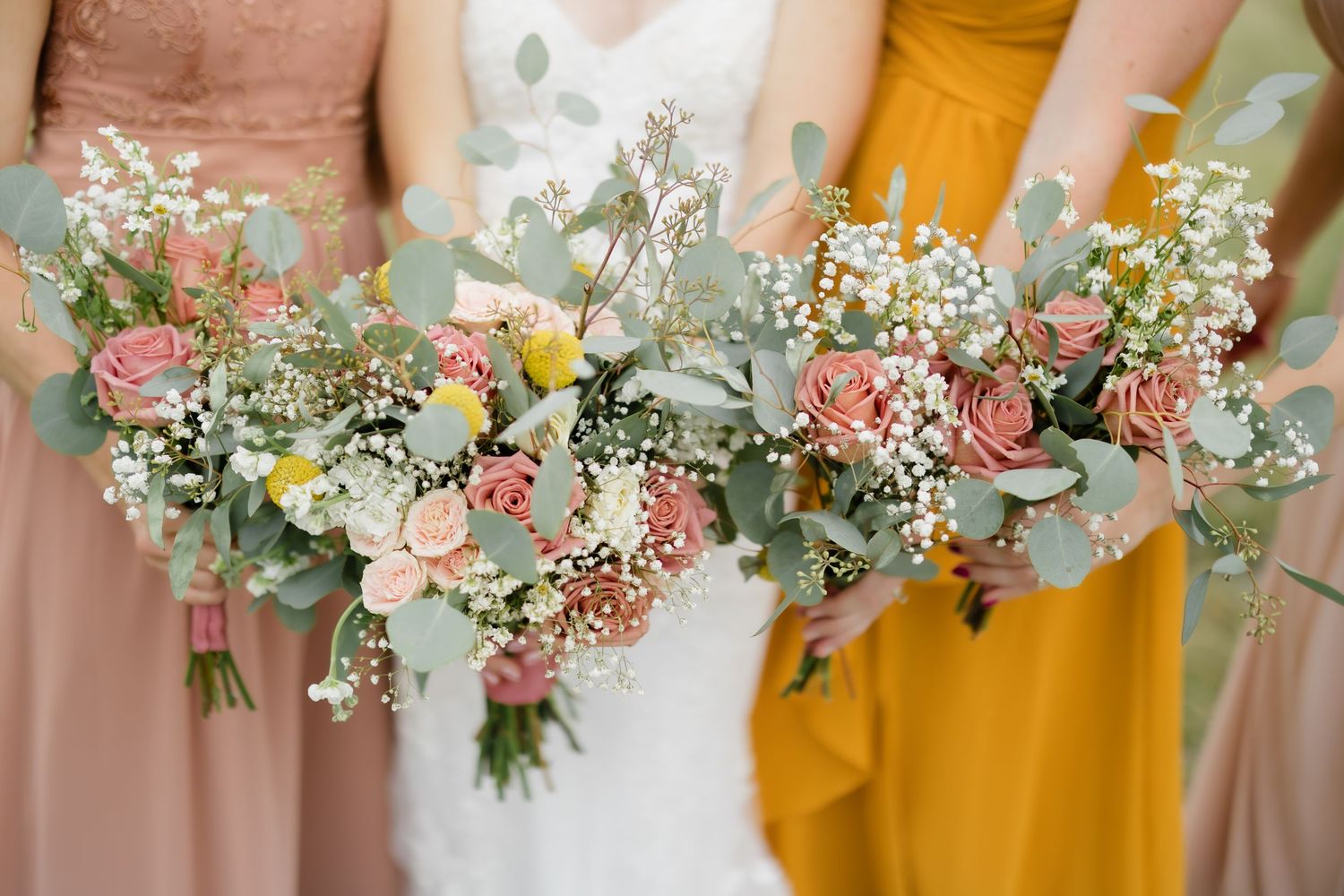 Bridesmaids in pink and mustard dresses holding bouquets of roses, eucalyptus, and baby's breath at a wedding.