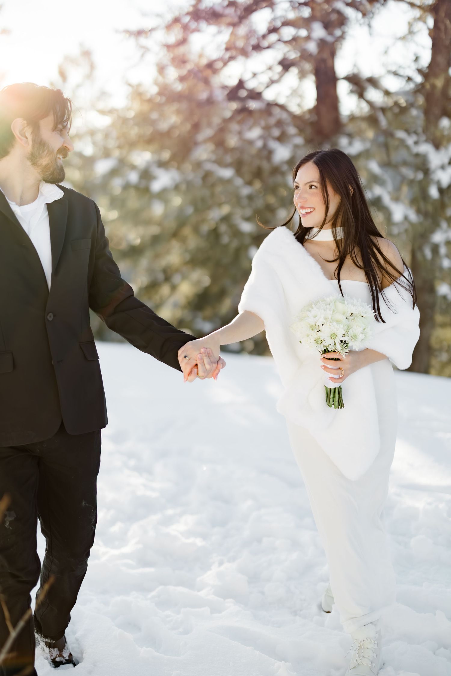 Bride and groom hold hands walking through snow-covered forest on their winter wedding day.