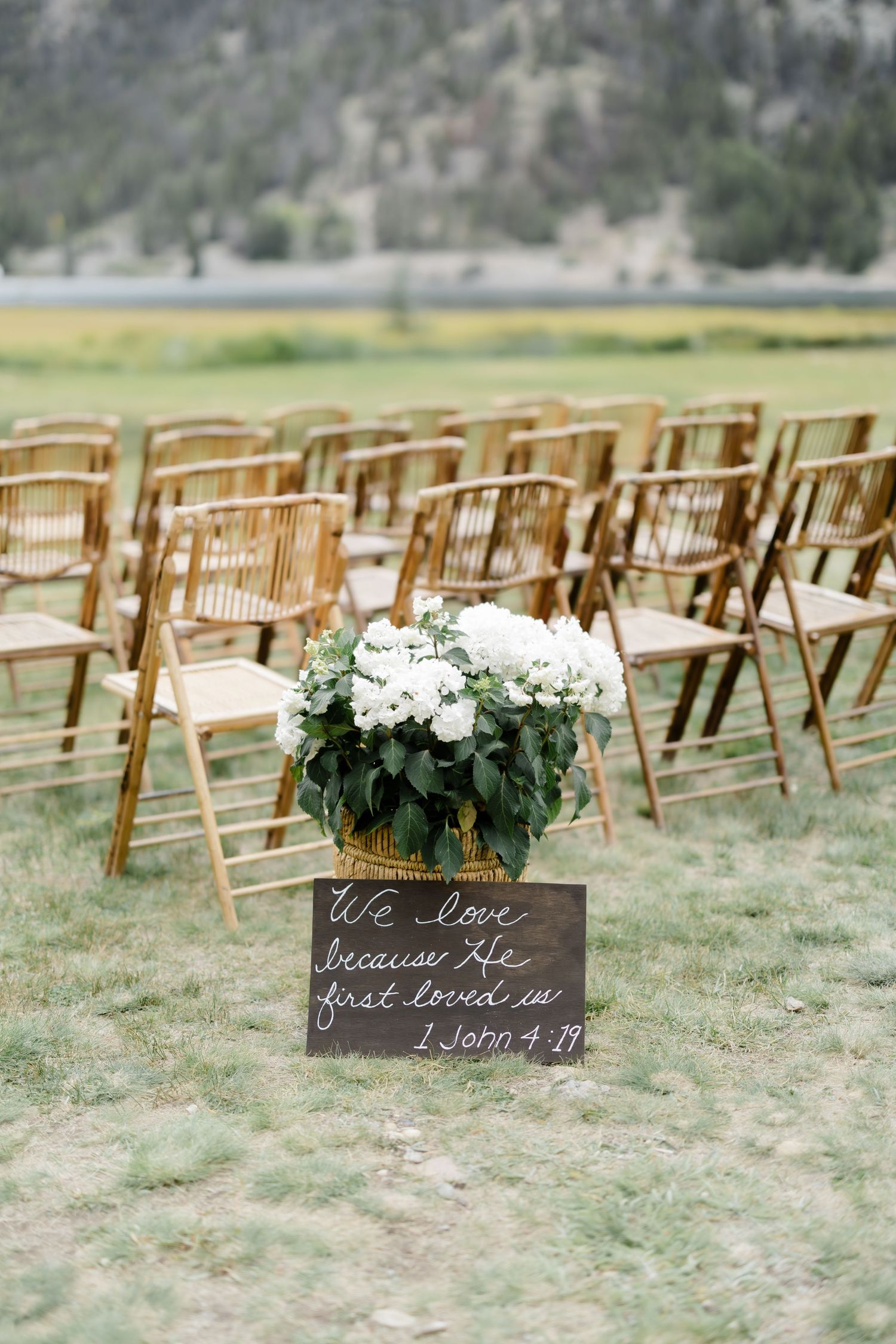 Wooden folding chairs arranged outdoors for a wedding ceremony with white hydrangeas and a scripture sign reading 1 John 4:19.