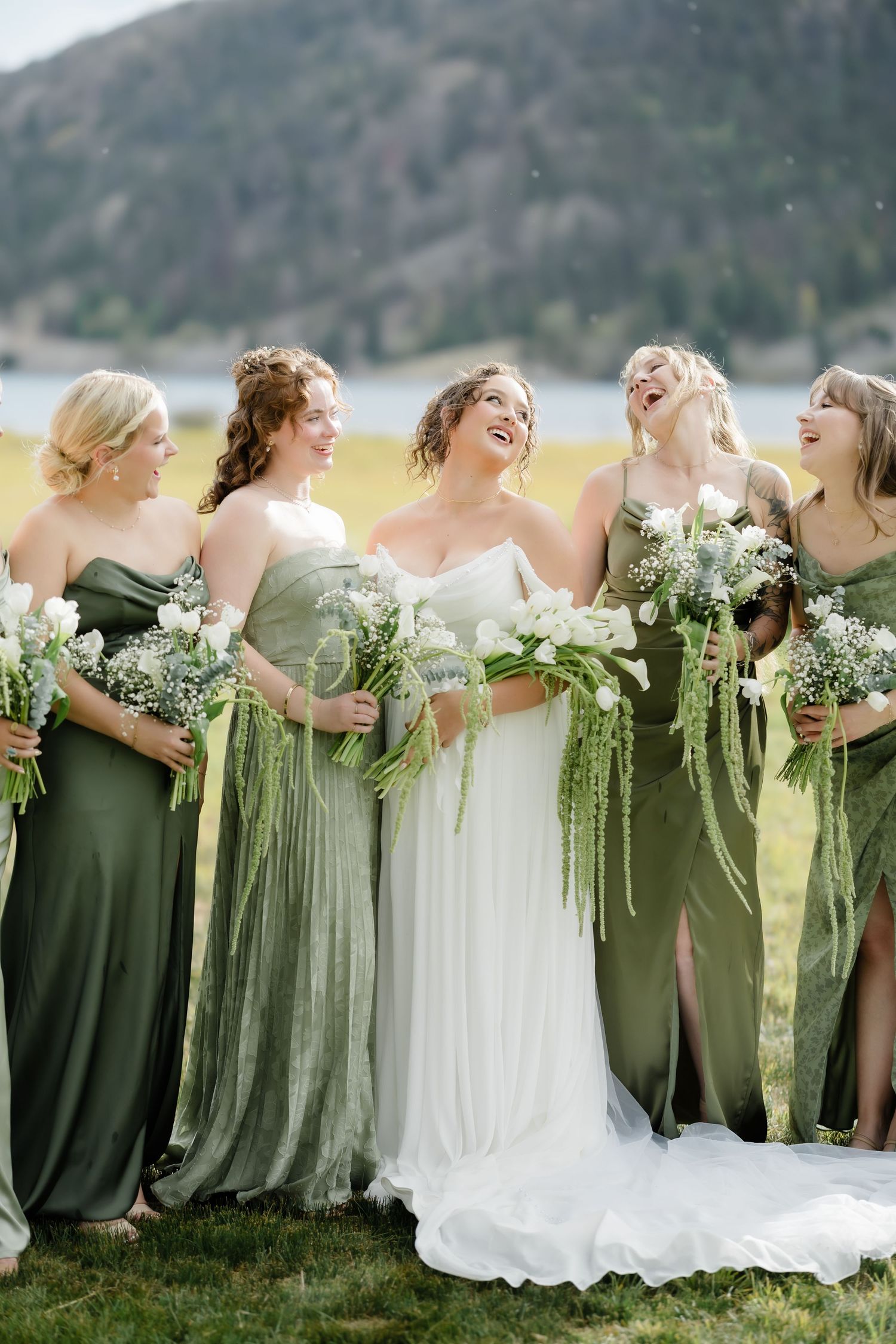 Bride in white gown laughs with bridesmaids in mismatched green dresses holding floral bouquets outdoors.