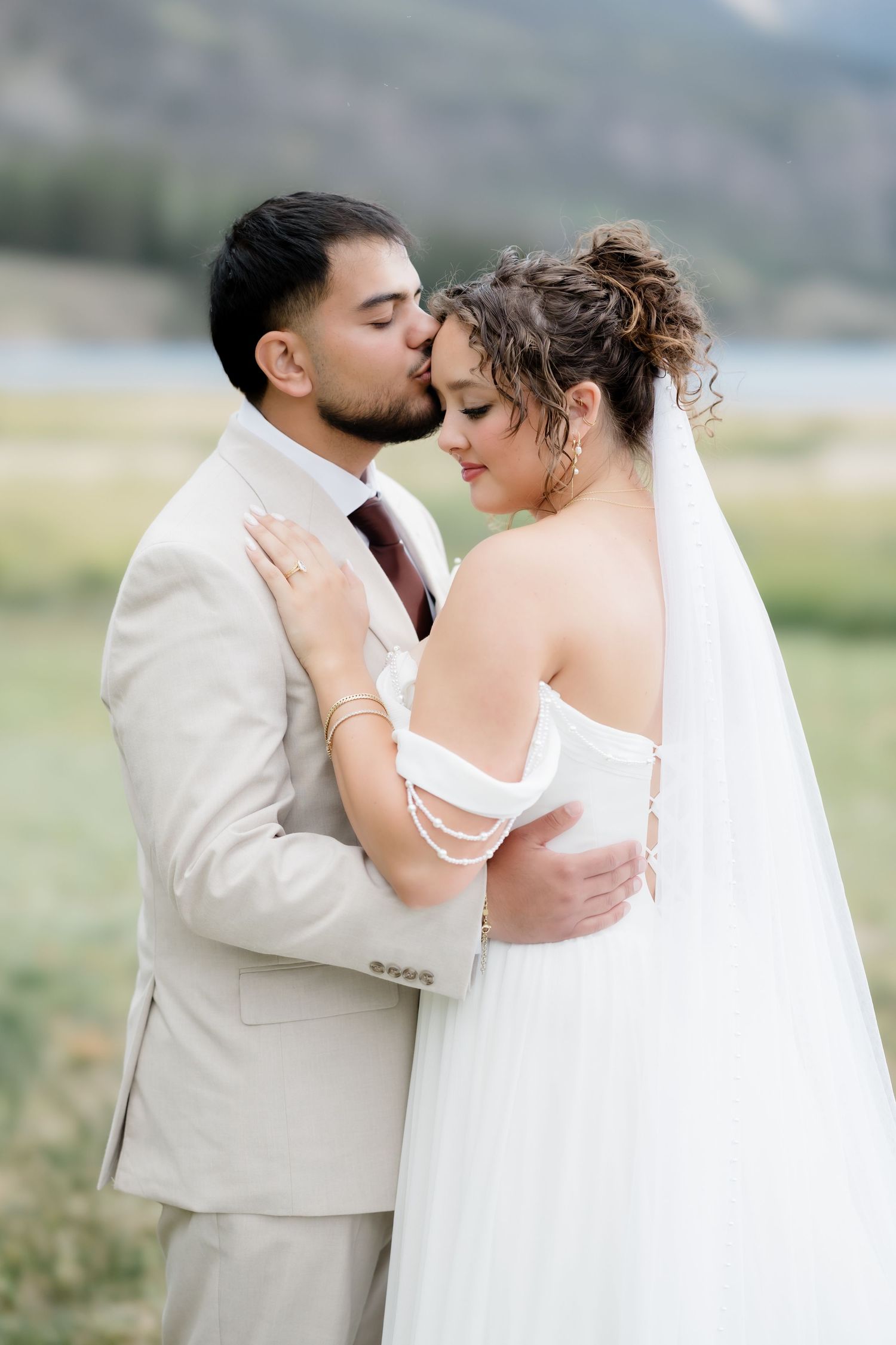 Groom in beige suit kisses bride in white off-shoulder gown outdoors near a lake with mountains in background.