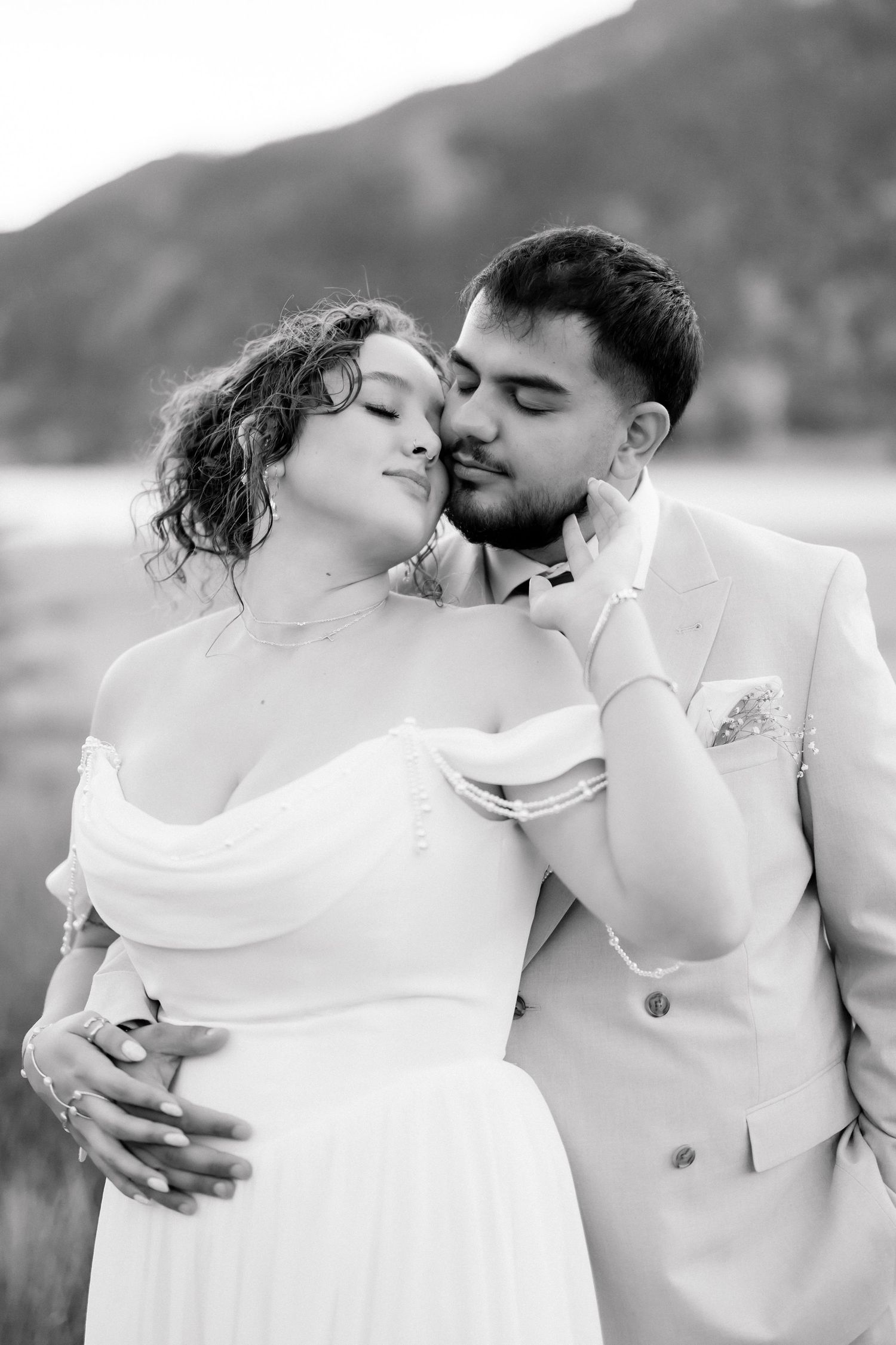 Black and white photo of a couple in wedding attire embracing outdoors with mountains in the background.