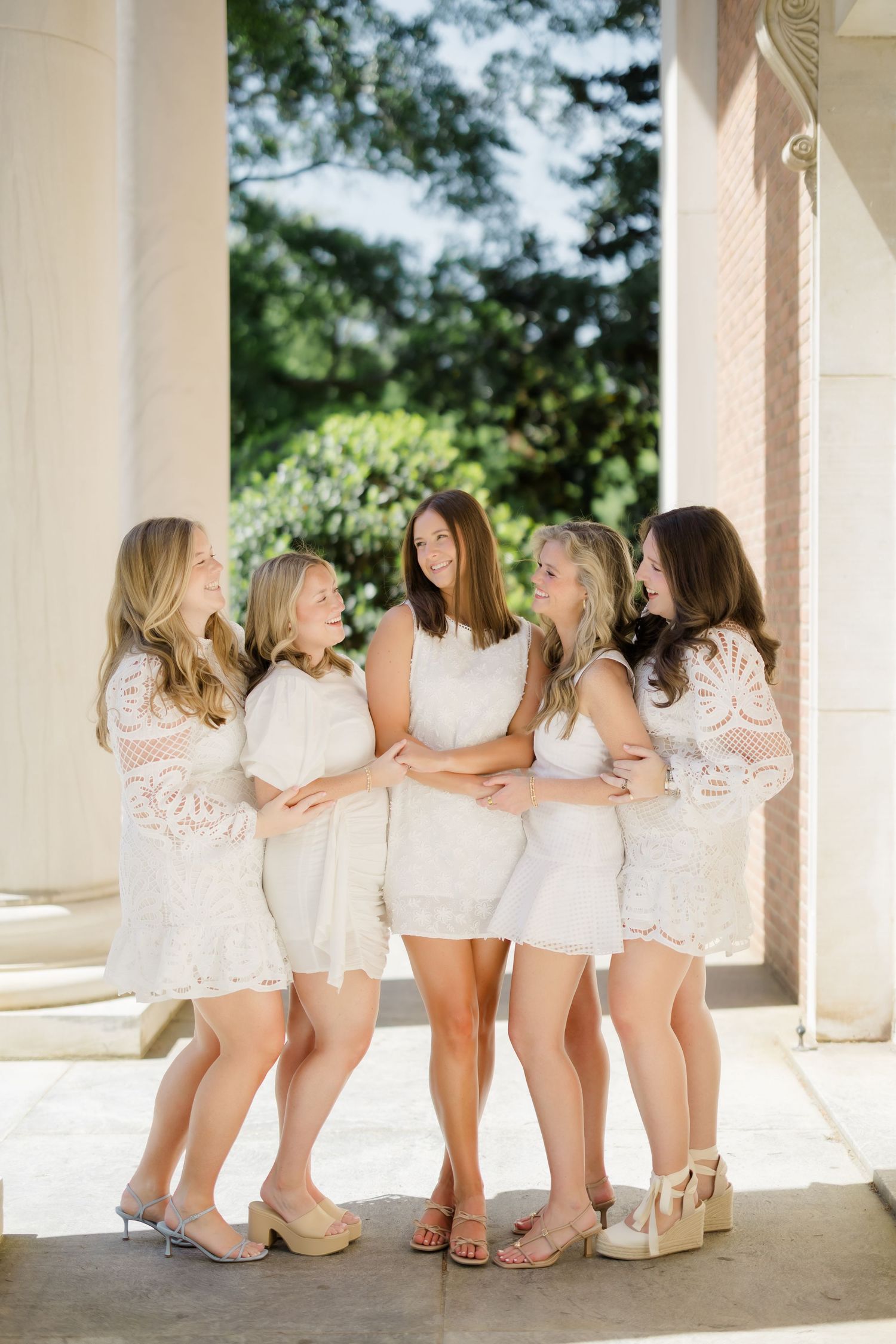 Five women in white lace dresses pose together laughing between columns outdoors.