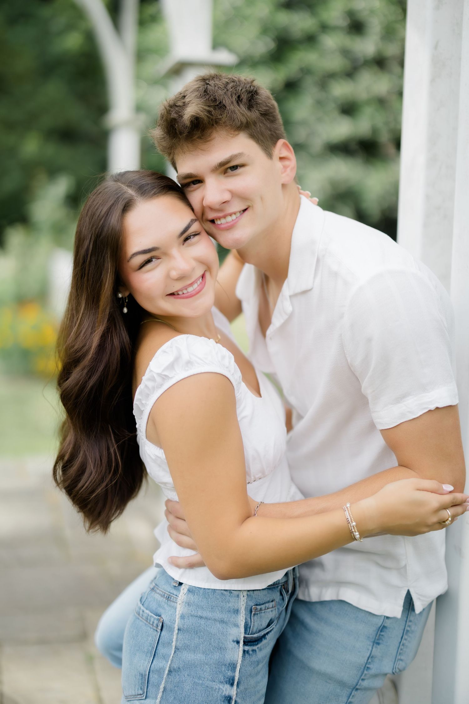 A smiling young couple in white tops and jeans embrace outdoors, leaning against a white pillar with greenery behind them.