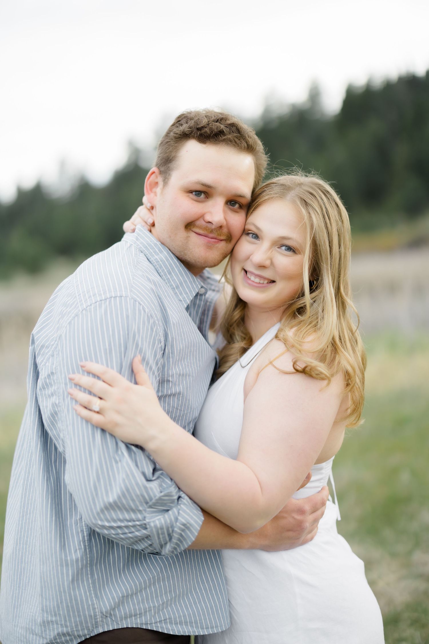 A smiling couple embraces outdoors, woman in white dress and man in blue striped shirt, with green trees in background.