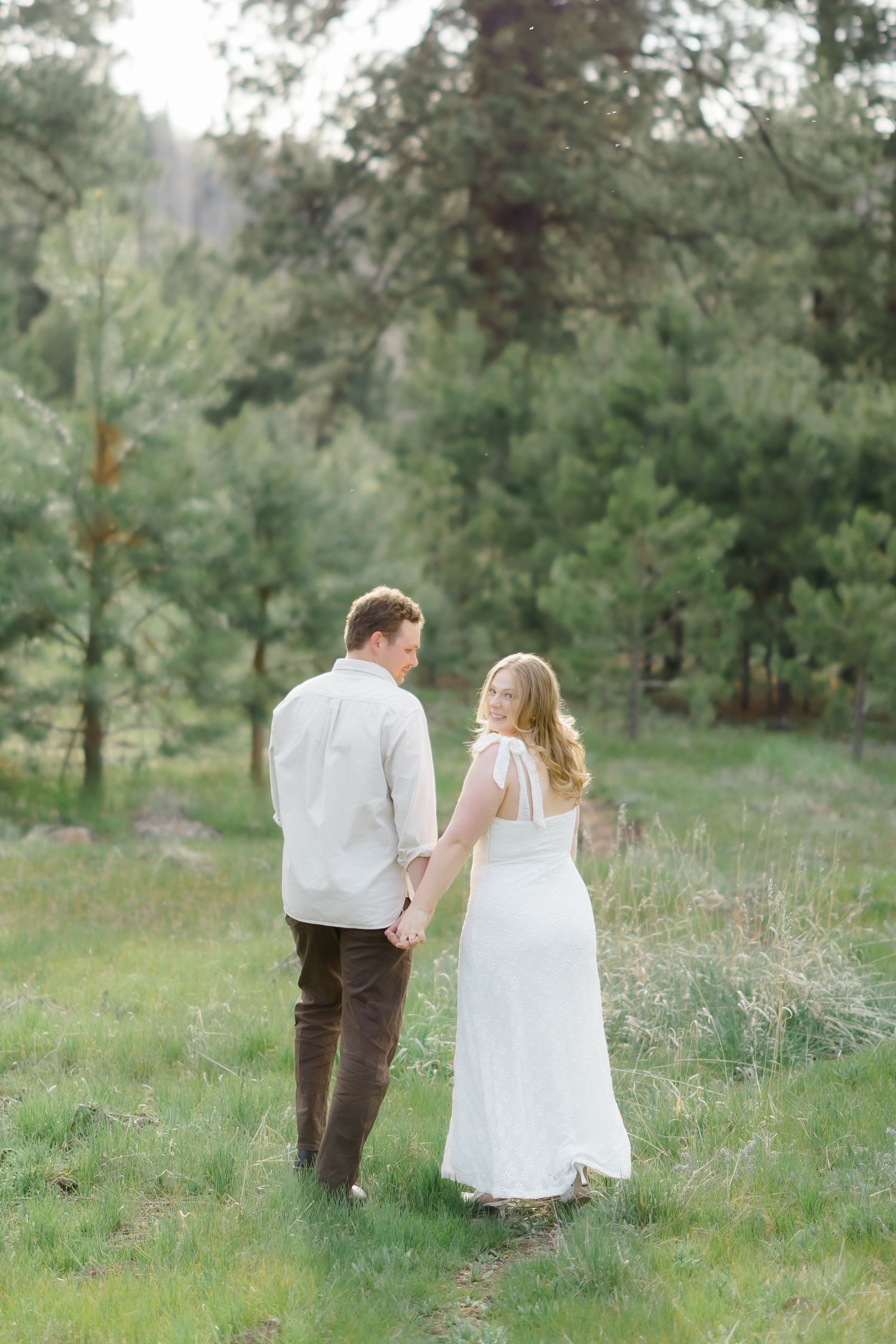 Couple holding hands walking through a lush green forest meadow, woman in white dress glancing back over shoulder.