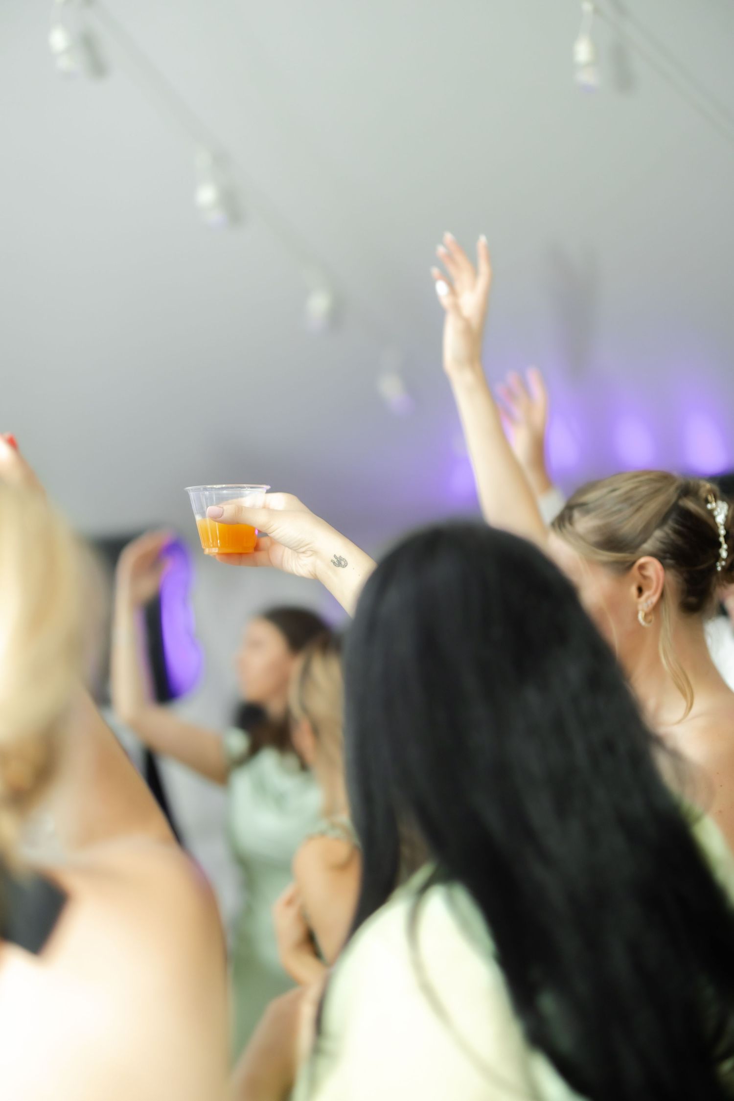 Wedding guests dance and raise hands at a lively reception, one holding an orange drink under string lights.