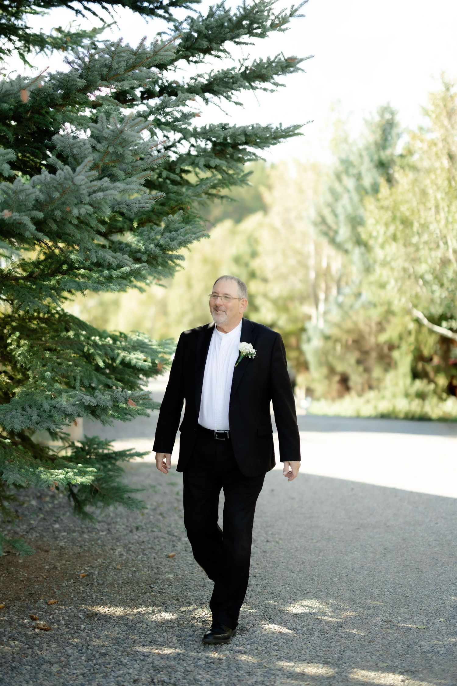 A well-dressed groom in a black suit with boutonniere walks along a gravel path surrounded by lush green trees.