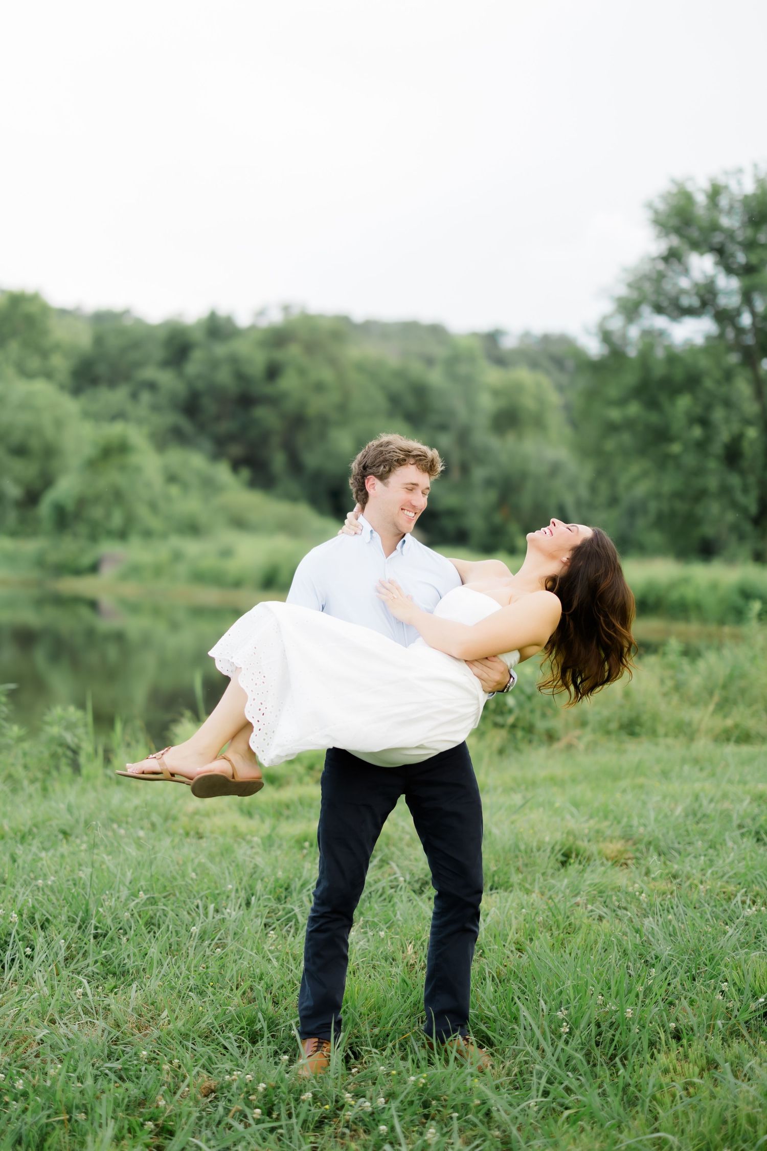 Man lifts woman in white dress outdoors by a pond, both smiling joyfully in a lush green meadow.