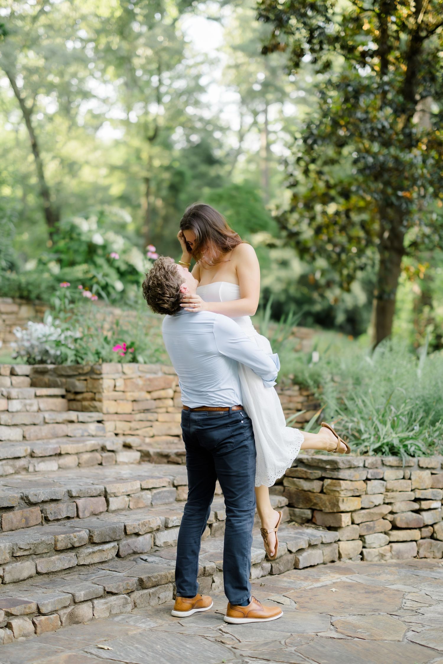 A man lifts a woman in a white dress in a lush garden with stone steps during a romantic engagement session.
