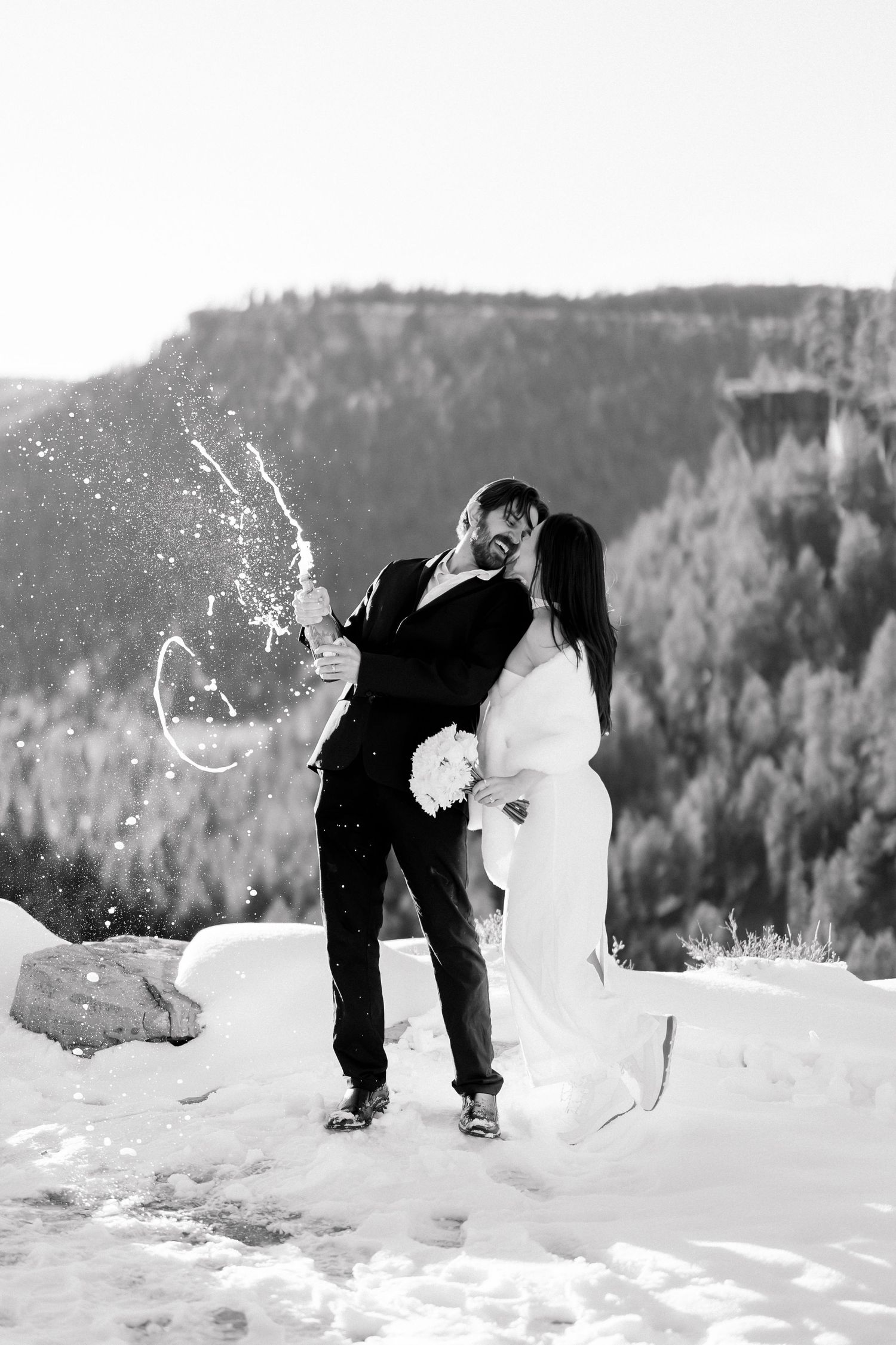Bride and groom kiss while popping champagne in snowy mountain setting, black and white wedding photo.
