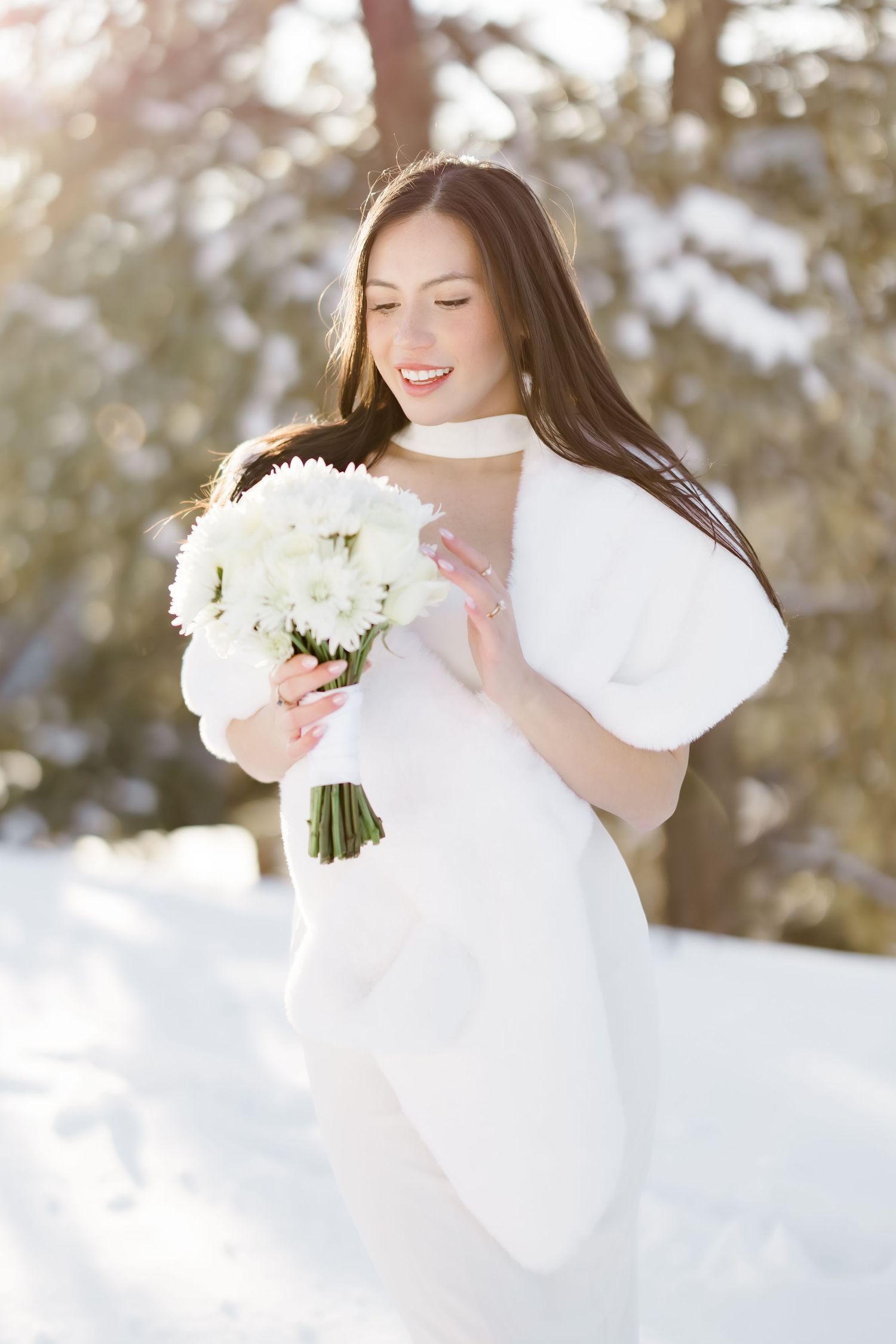 Radiant bride in white gown and fur wrap holds white flower bouquet in snowy winter setting.
