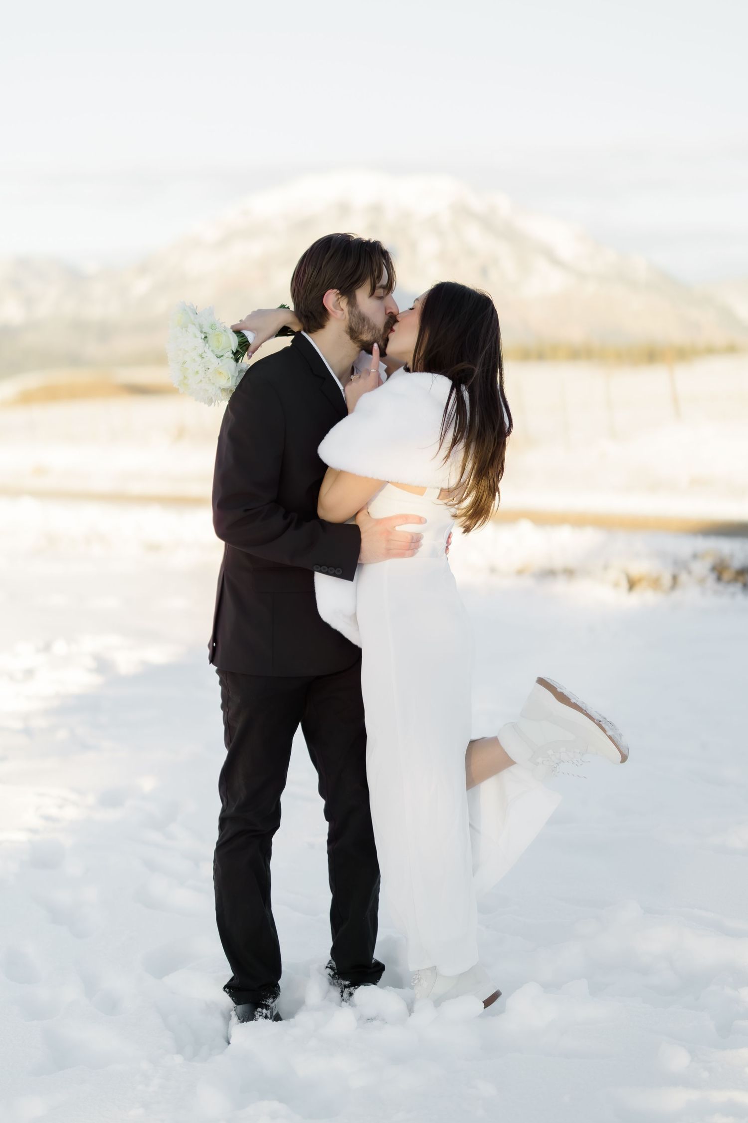 Bride and groom share romantic kiss in snow with mountain backdrop on wedding day.