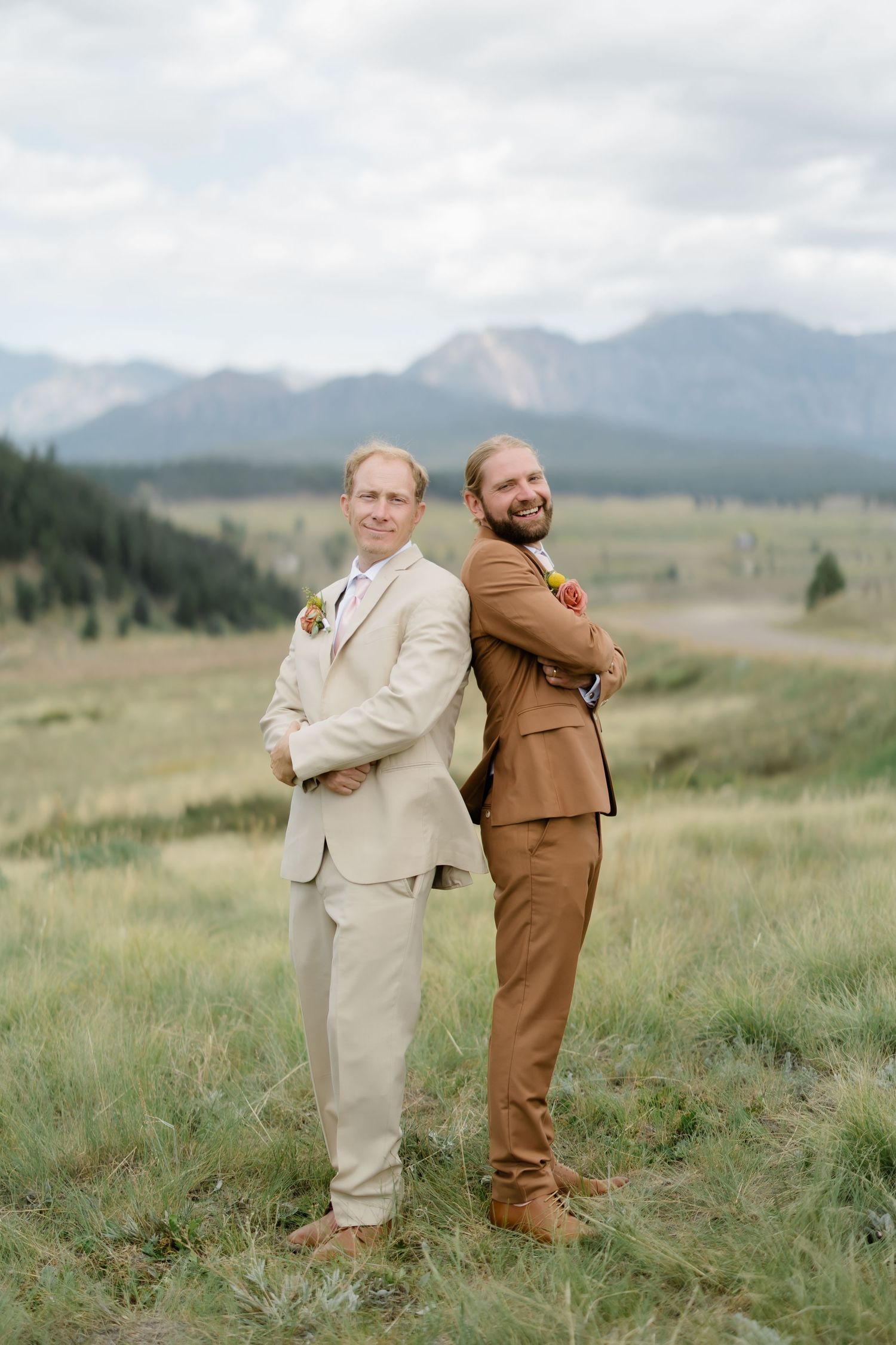 Two grooms in tan and brown suits stand back-to-back smiling in a mountain meadow on their wedding day.