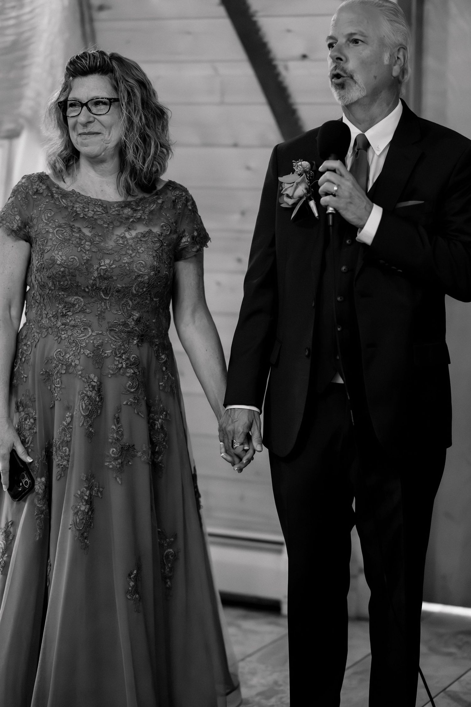 Black and white photo of an elegant couple holding hands at a wedding, man speaking into microphone wearing a suit with boutonniere.